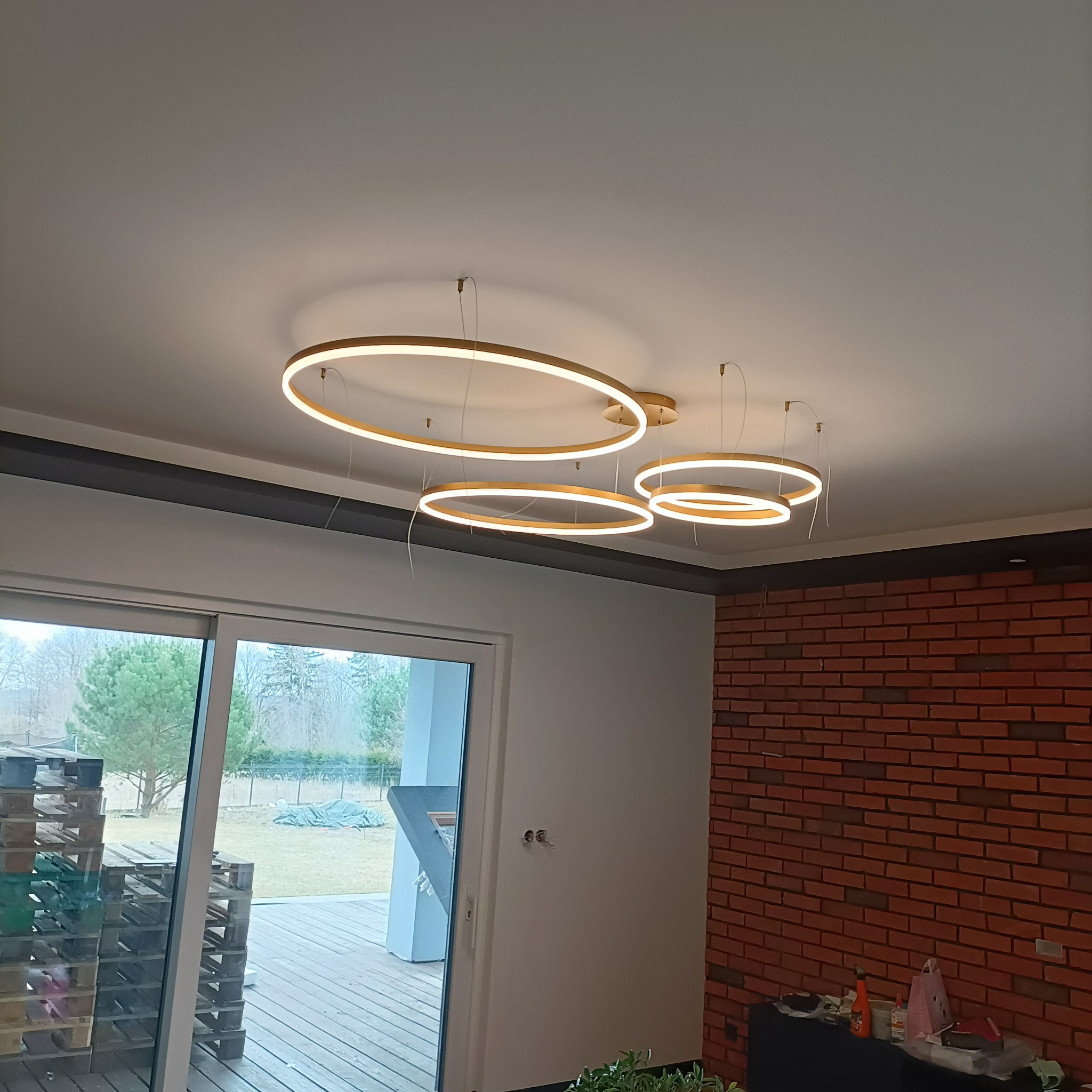 Nowoczesne oświetlenie sufitowe LED w kształcie pierścieni, zawieszone w jasnym wnętrzu z ceglaną ścianą i dużym oknem.