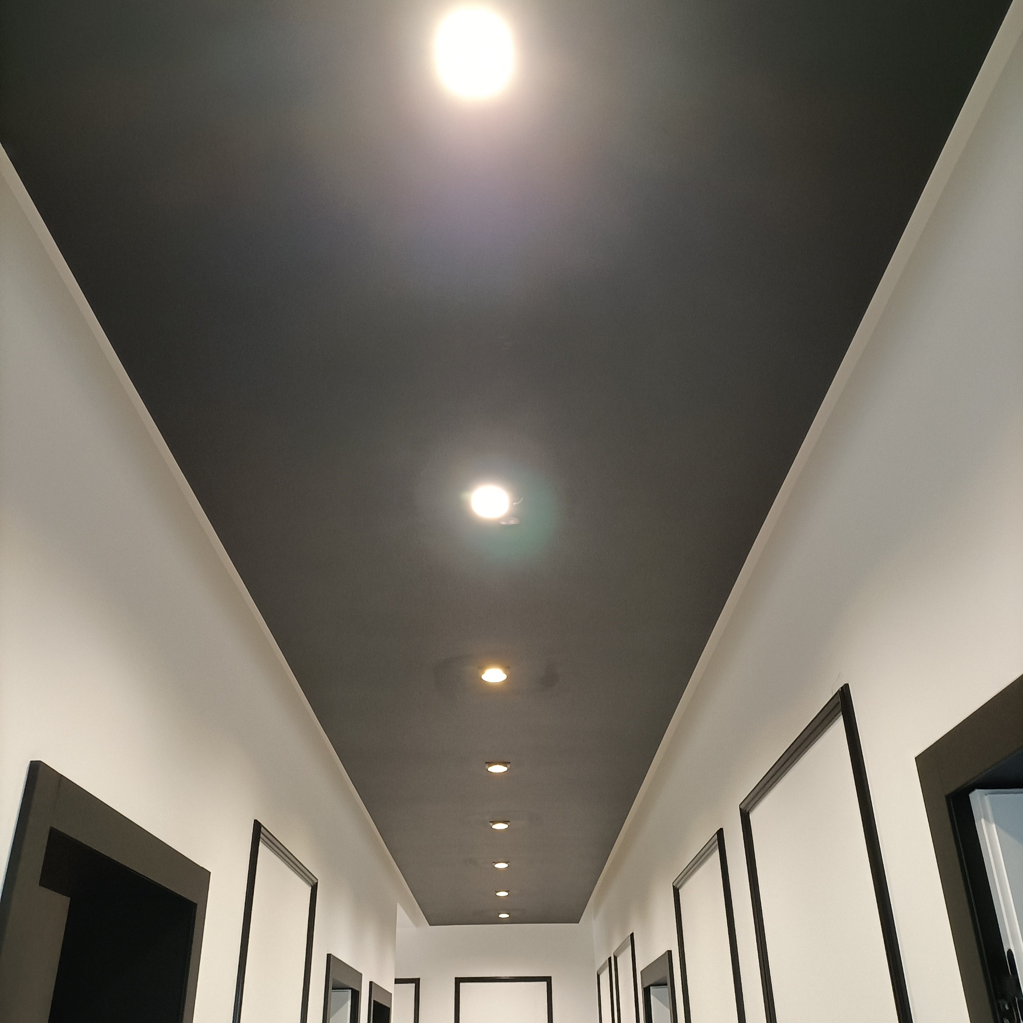 Długi korytarz z czarnym sufitem i rzędem punktowych lamp. Białe ściany z czarnymi obramowaniami drzwi. Perspektywa zbieżna ku końcowi korytarza.