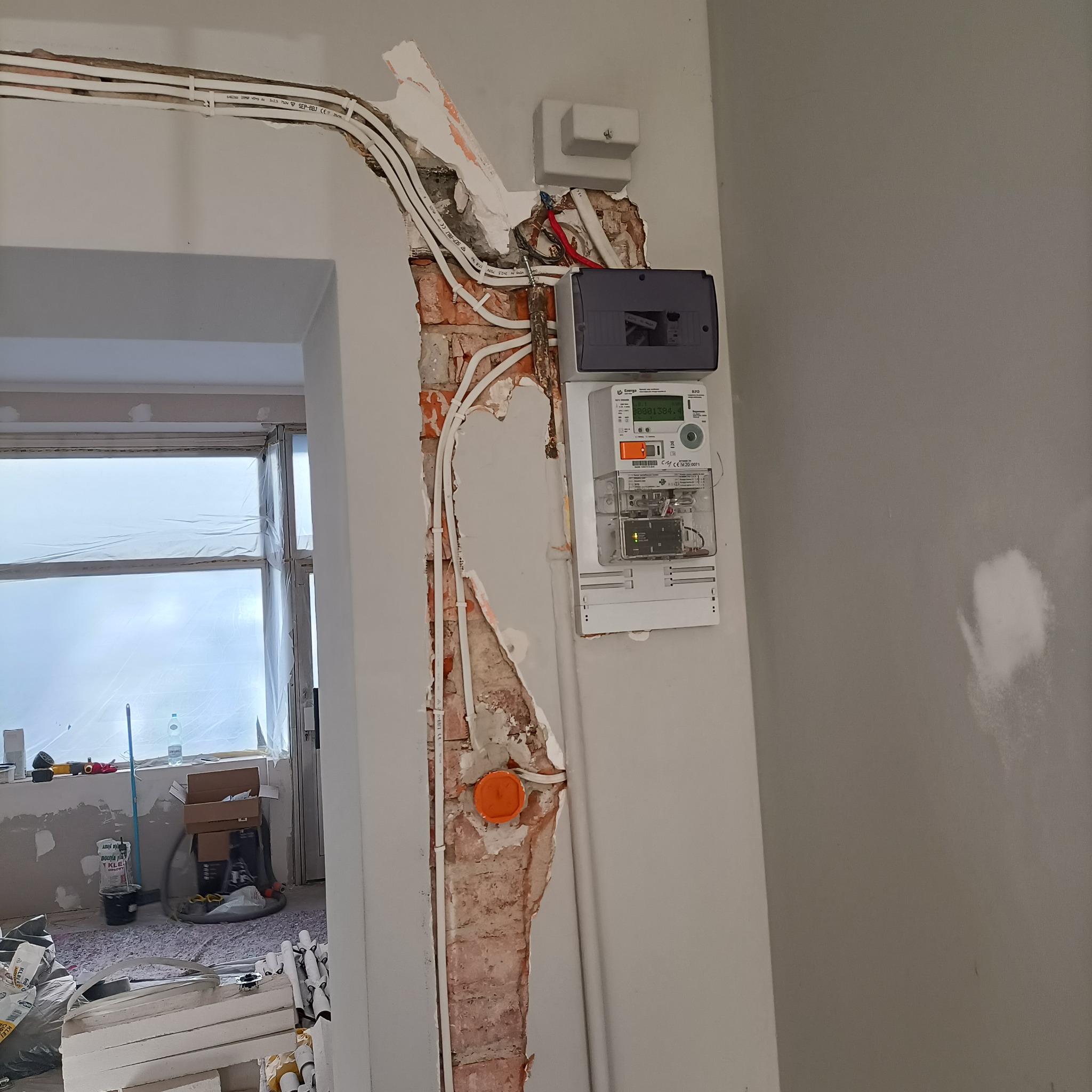 Fragment ściany z odkrytą instalacją elektryczną, widoczne przewody w peszlach, licznik i puszka. W tle pomieszczenie w trakcie remontu.