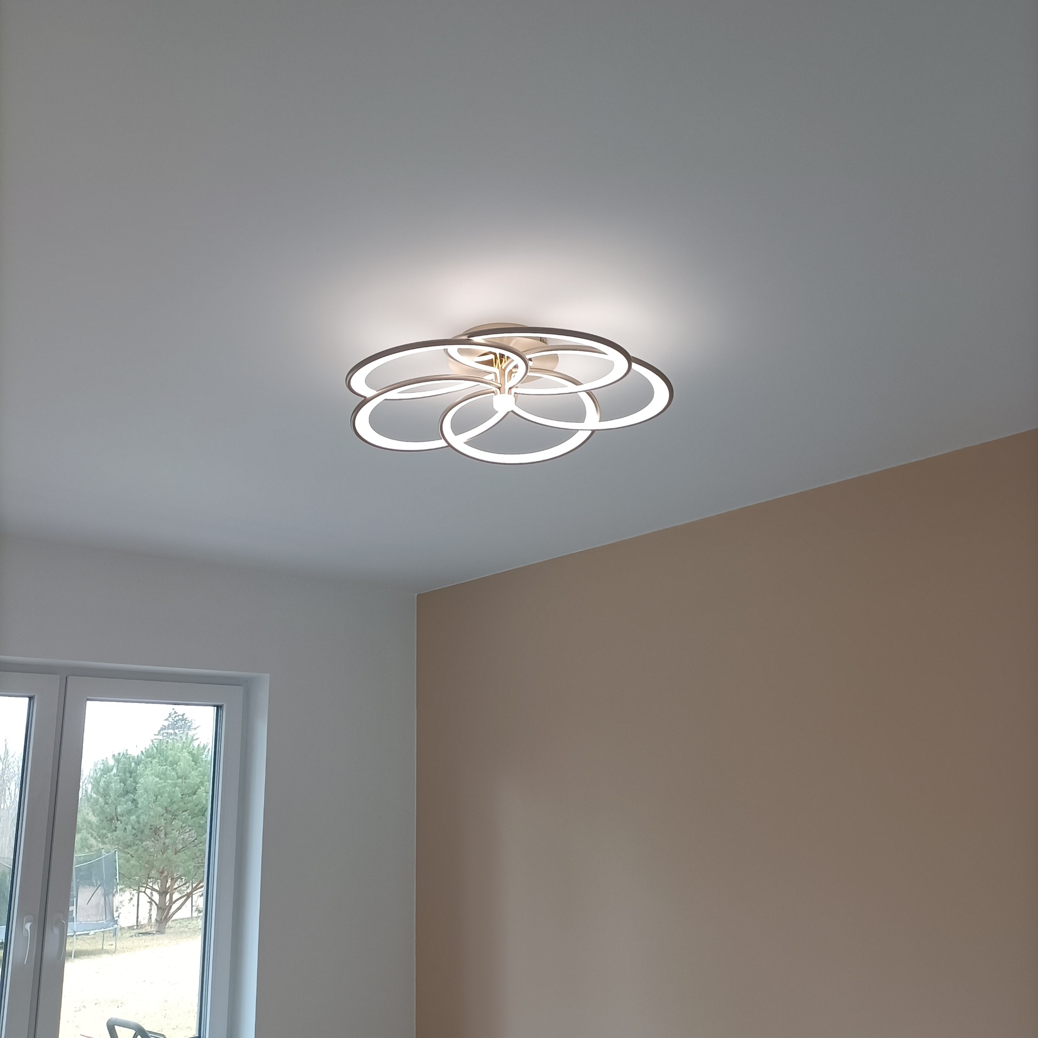 Nowoczesna lampa sufitowa LED w kształcie kwiatu z okrągłych pierścieni, zamontowana w jasnym pomieszczeniu z oknem. Neutralne barwy ścian.