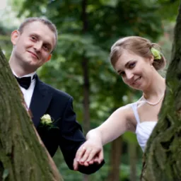 Para młoda trzymająca się za ręce, spoglądająca zza drzew na tle zieleni. Pan młody w smokingu, panna młoda w białej sukni i perłach. Ujęcie z bliska, naturalne światło.