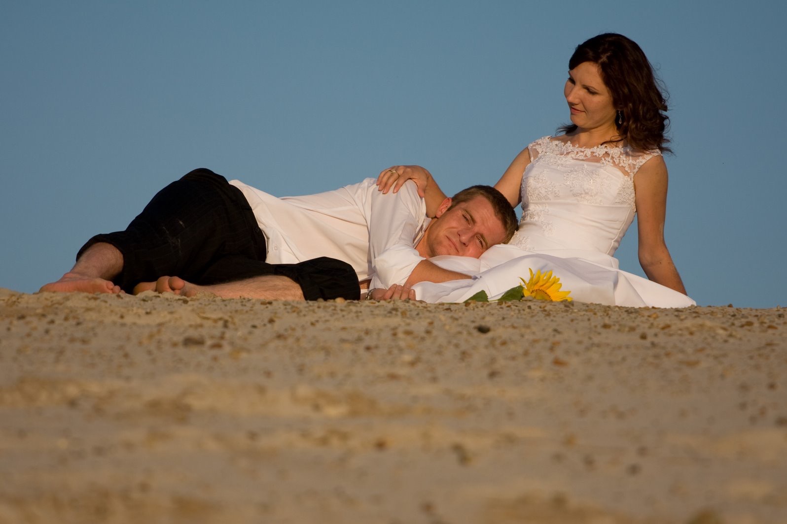 Para młoda leżąca na piasku z słonecznikiem; on w białej koszuli i czarnych spodniach, ona w białej sukni ślubnej, uśmiechnięta, z pierścionkiem na palcu.