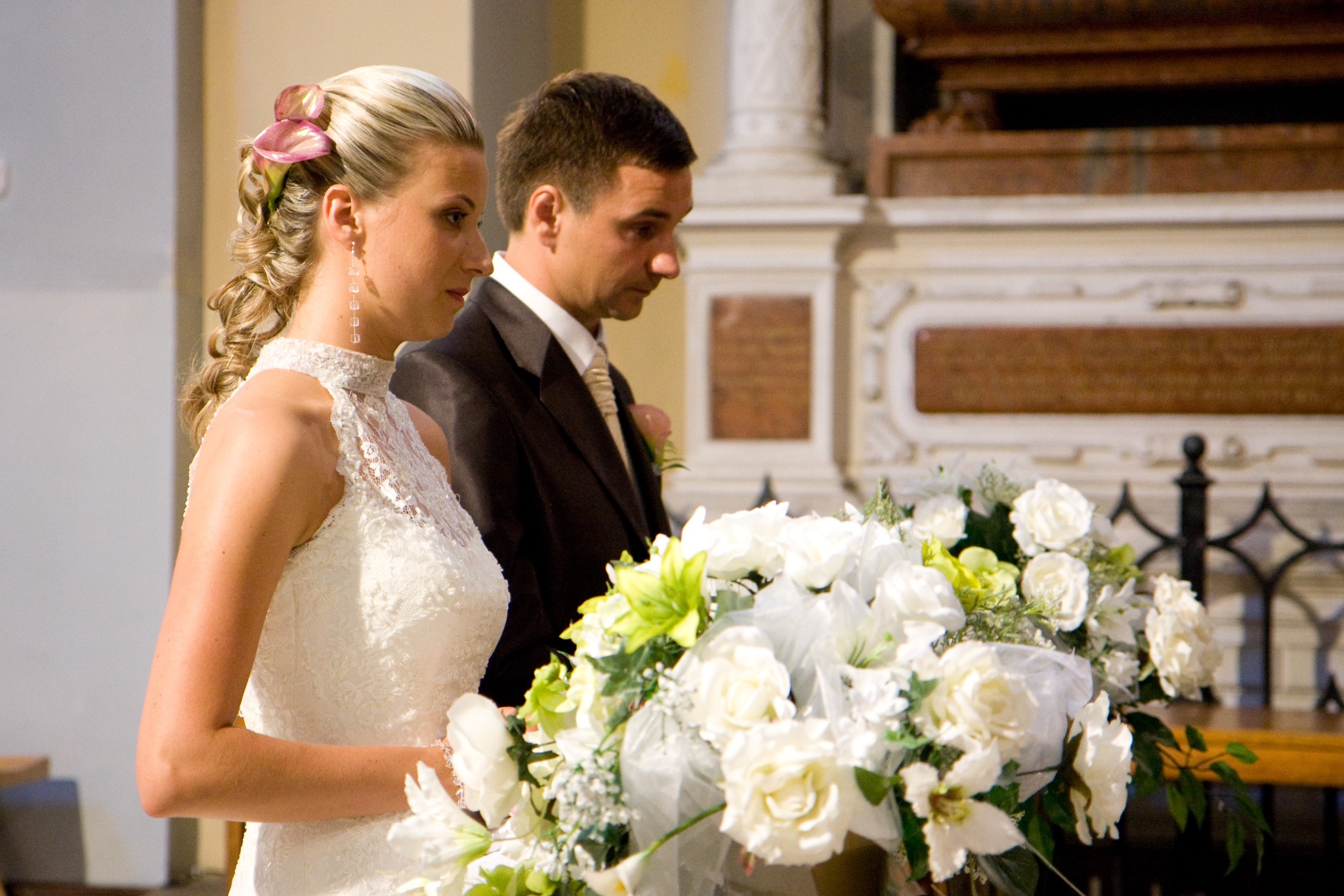 Para młoda podczas ceremonii ślubnej w kościele. Panna młoda w sukni z koronką, pan młody w ciemnym garniturze. Bukiet białych róż na pierwszym planie, ozdobny ołtarz w tle.