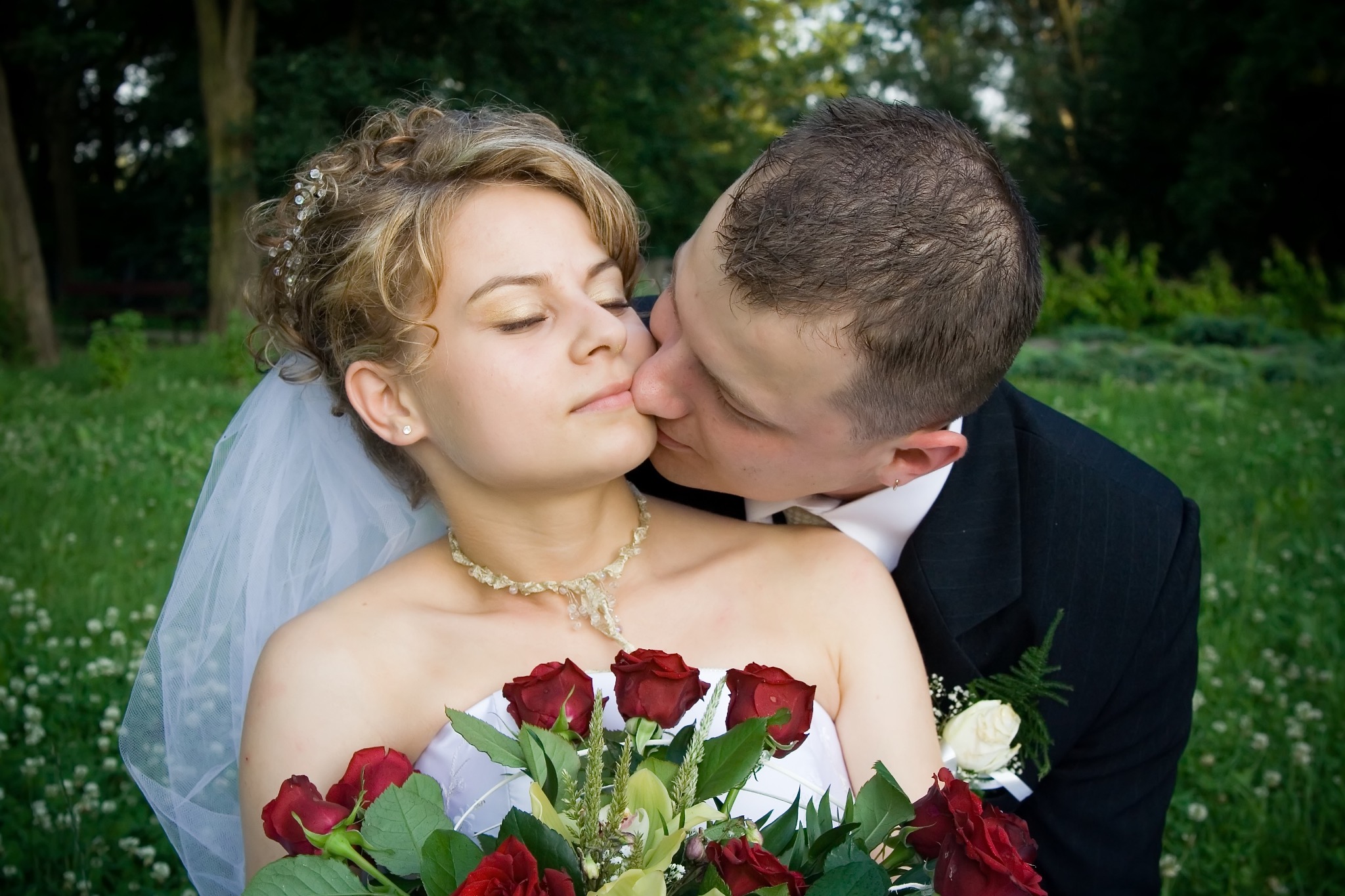 Pocałunek nowożeńców w plenerze: panna młoda z bukietem czerwonych róż i welonem, pan młody w czarnym garniturze. Romantyczna chwila w zielonym otoczeniu.