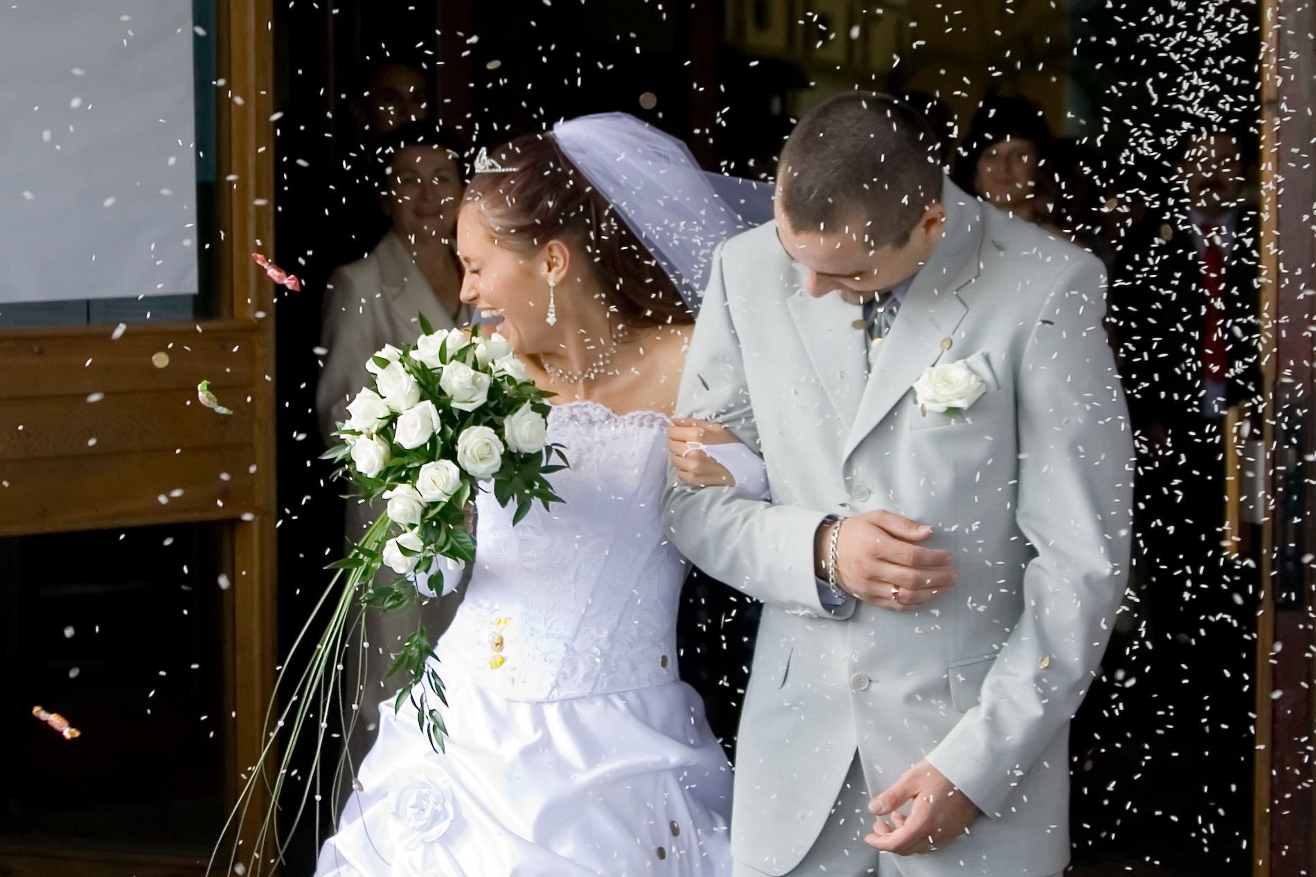 Uśmiechnięta panna młoda z bukietem i pan młody w szarym garniturze wychodzą z kościoła w deszczu konfetti. W tle goście.