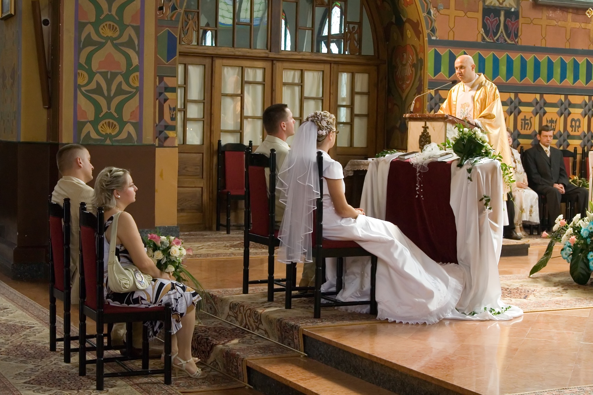 Uroczystość ślubna w Tarnobrzegu: Panna młoda w białej sukni siedzi w kościele podczas ceremonii, obok goście, w tle ksiądz przy ołtarzu i bogato zdobione ściany.