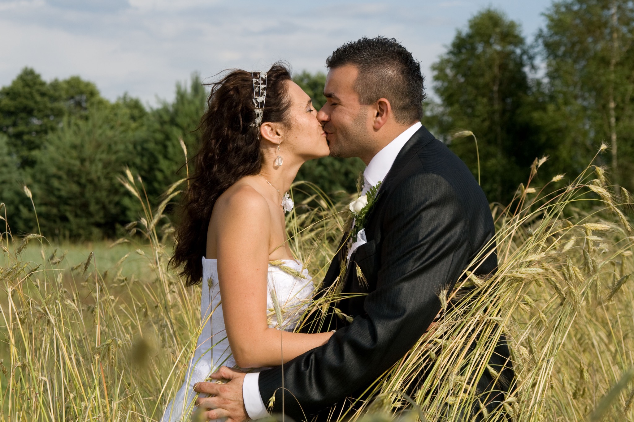 Pocałunek młodej pary w plenerze, panna młoda w białej sukni z ozdobą we włosach, pan młody w czarnym garniturze, tło stanowią drzewa i pole pszenicy.