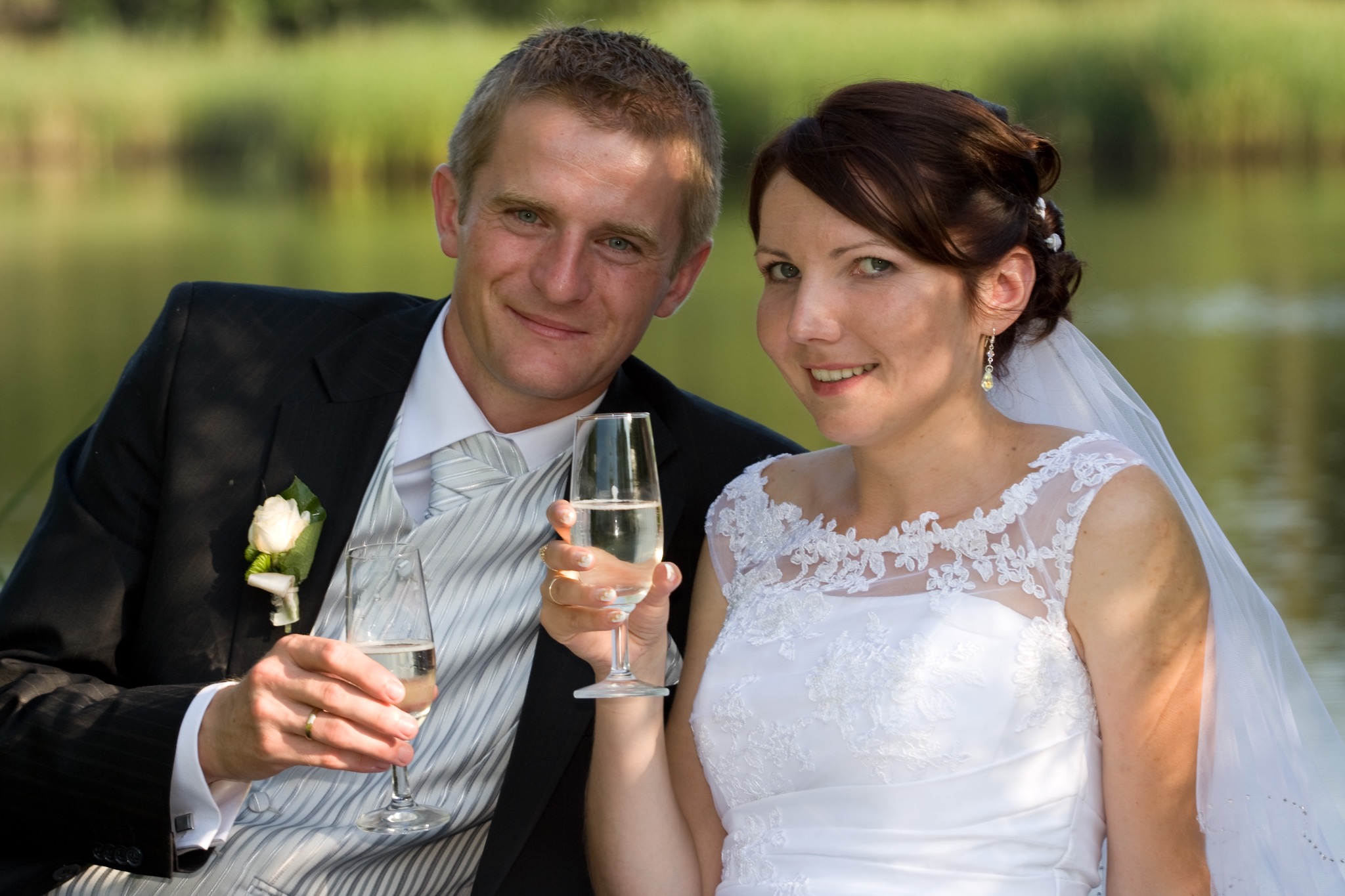 Uśmiechnięta para młoda w plenerze, trzymająca kieliszki z szampanem. Pan w czarnym garniturze, pani w białej sukni ślubnej z koronkowym wykończeniem.