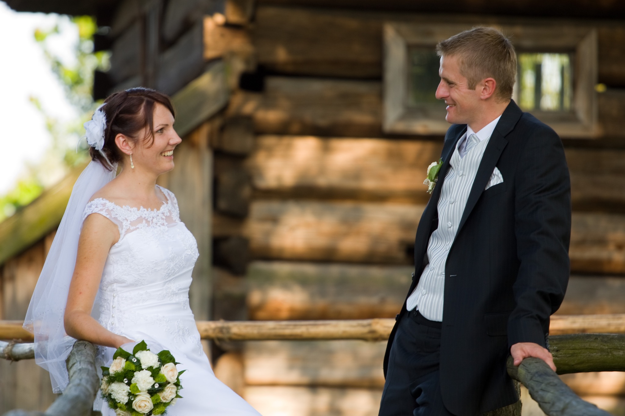 Uśmiechnięta para młoda w plenerze, ona w białej sukni z bukietem, on w eleganckim garniturze, oboje opierają się o drewnianą barierkę na tle drewnianego budynku.