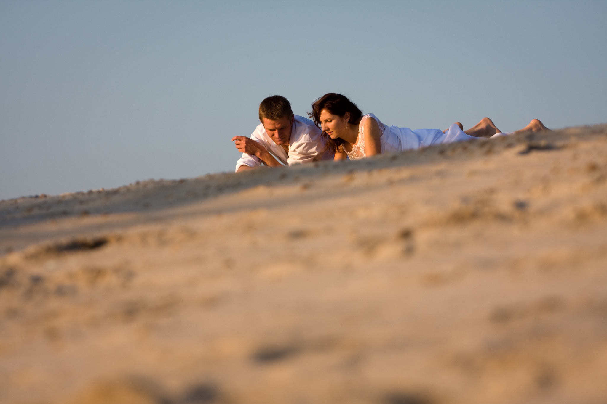 Para młoda leży na piaszczystej wydmie w białych strojach. Delikatne oświetlenie podkreśla romantyczny nastrój sesji plenerowej. Horyzont delikatnie rozmyty.