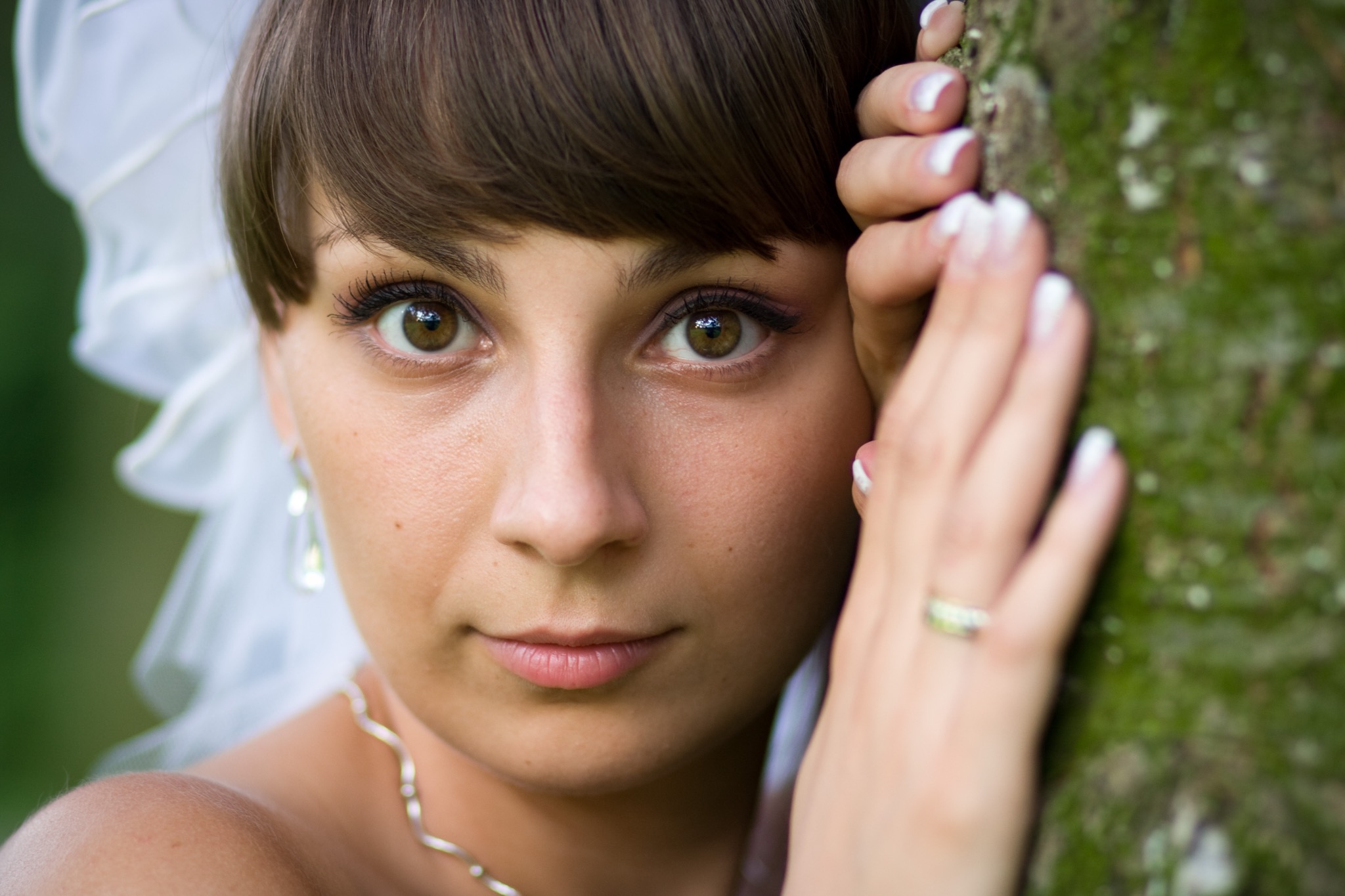 Portret panny młodej z brązowymi oczami, opierającej się o drzewo. Widoczne delikatne kolczyki, naszyjnik i welon. Makijaż podkreśla naturalne piękno.