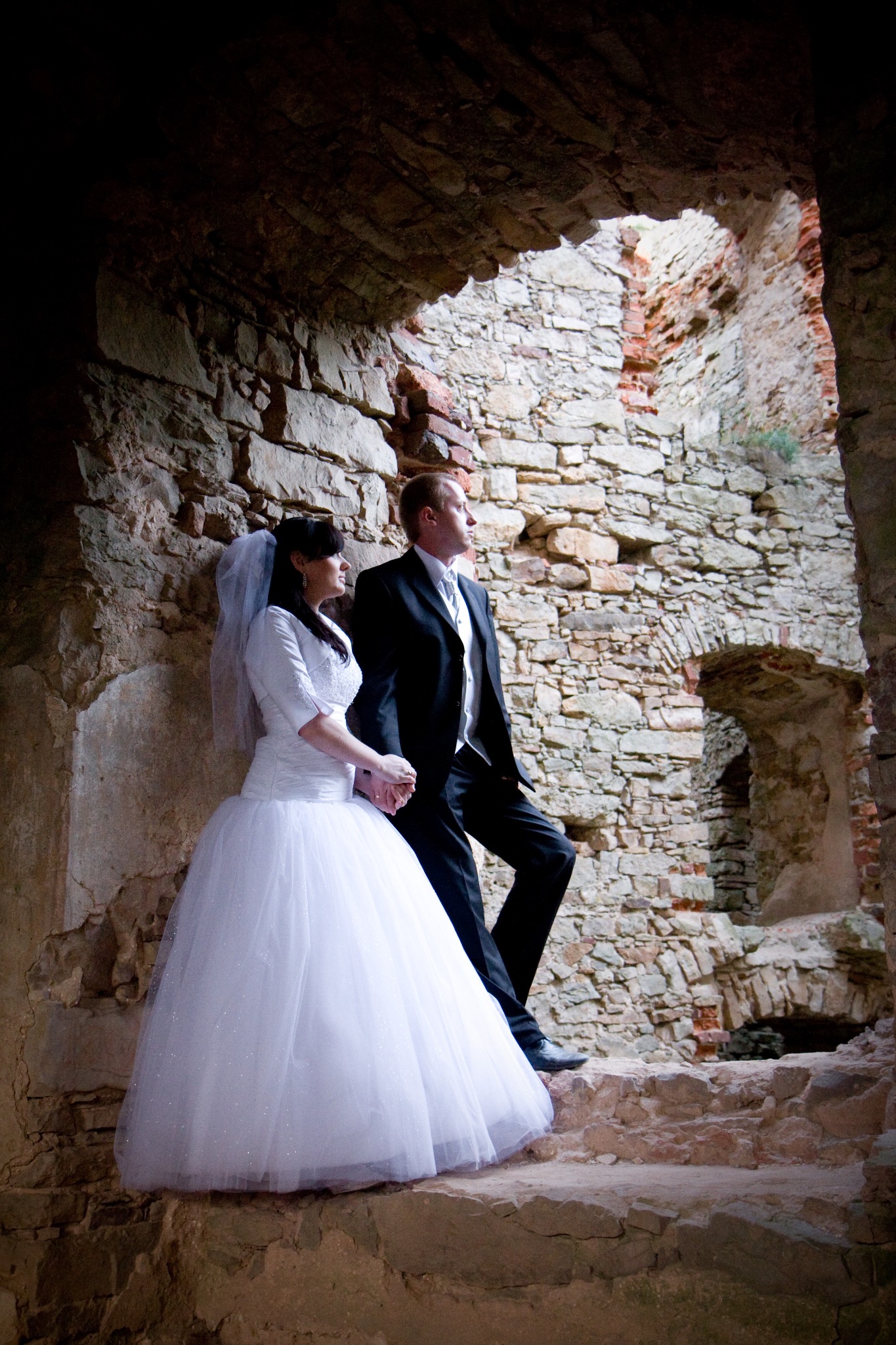 Para młoda w ruinach zamku: panna młoda w białej sukni i welonie, pan młody w czarnym garniturze, trzymają się za ręce na tle kamiennych murów.
