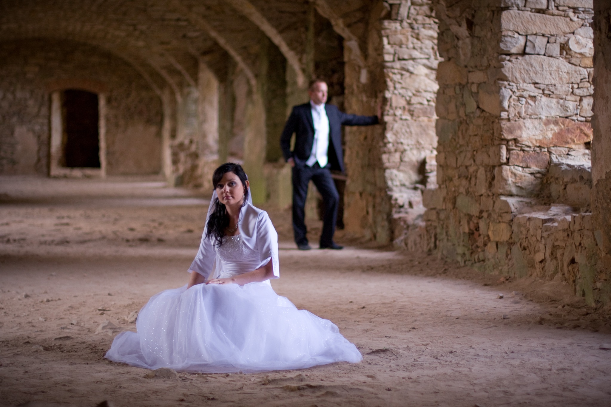 Para młoda: panna młoda siedzi na piasku w białej sukni, pan młody opiera się o kamienny filar w ruinach. Nietypowa sceneria ślubna.