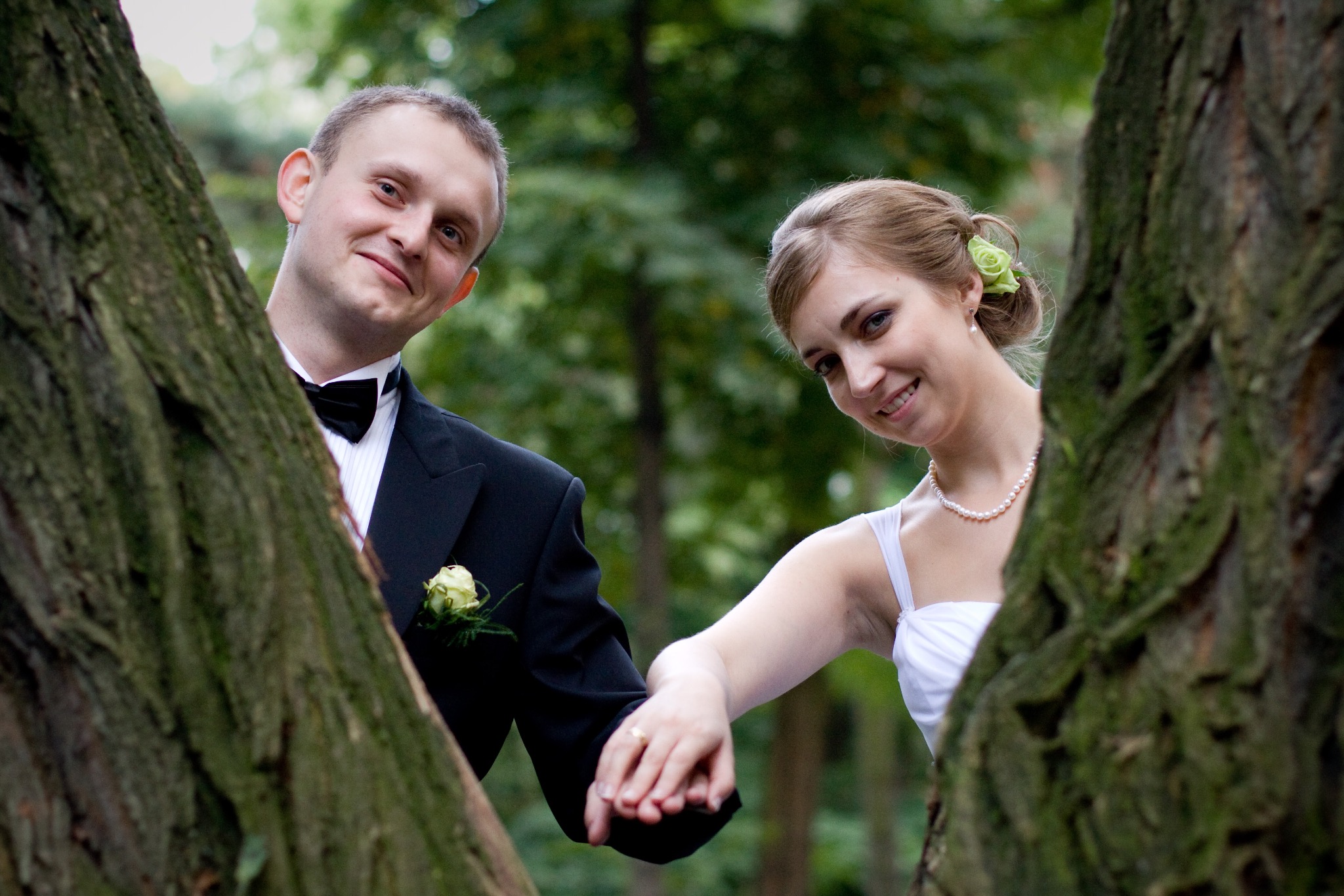 Para młoda trzymająca się za ręce, spoglądająca zza drzew na tle zieleni. Pan młody w smokingu, panna młoda w białej sukni i perłach. Ujęcie z bliska, naturalne światło.