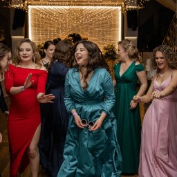 PHOTOLA Aleksandra Krzan - Grupa roześmianych kobiet w sukniach wieczorowych tańczy na parkiecie podczas imprezy, widoczne dekoracje świetlne i żyrandol.