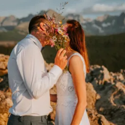 Para młoda całuje się za bukietem polnych kwiatów na tle górskiego krajobrazu o zachodzie słońca. Panna młoda w białej sukni z koronkowymi detalami, pan młody w białej koszuli i szarych spodniach.