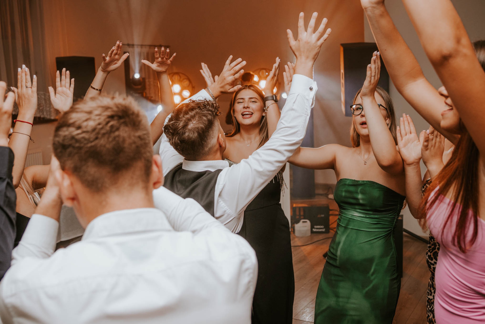 Rozbawieni goście z podniesionymi rękami podczas imprezy, widoczne damskie sukienki wieczorowe i męska koszula z kamizelką