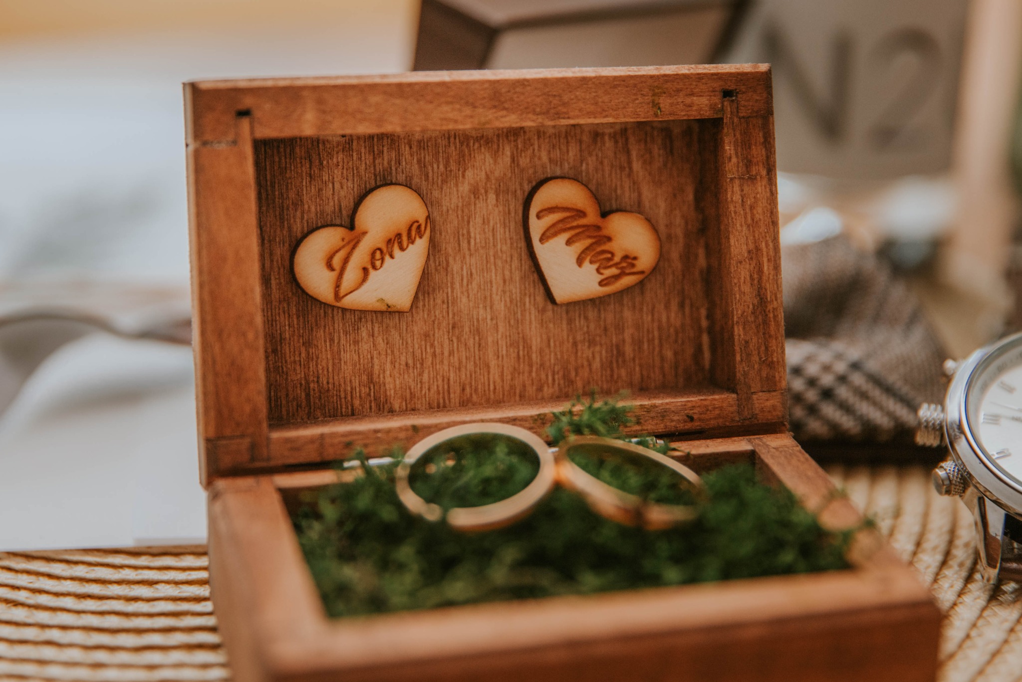 Drewniane pudełko na obrączki wypełnione mchem, z dekoracyjnymi sercami z napisami 'Żona' i 'Mąż', ustawione na plecionej macie obok zegarka.