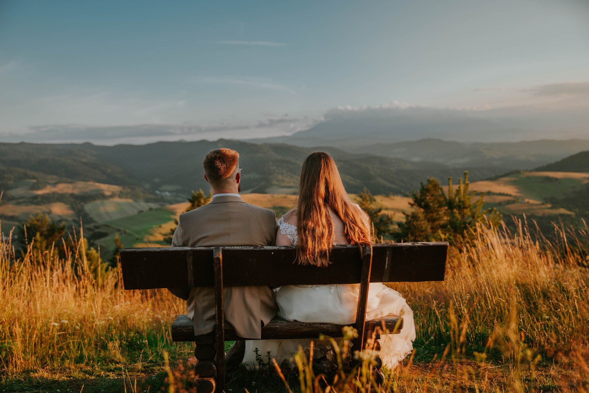 Para młoda siedzi na drewnianej ławce z widokiem na górzysty krajobraz o zachodzie słońca, panna młoda w białej sukni z koronkowymi detalami na plecach, pan młody w beżowym garniturze.