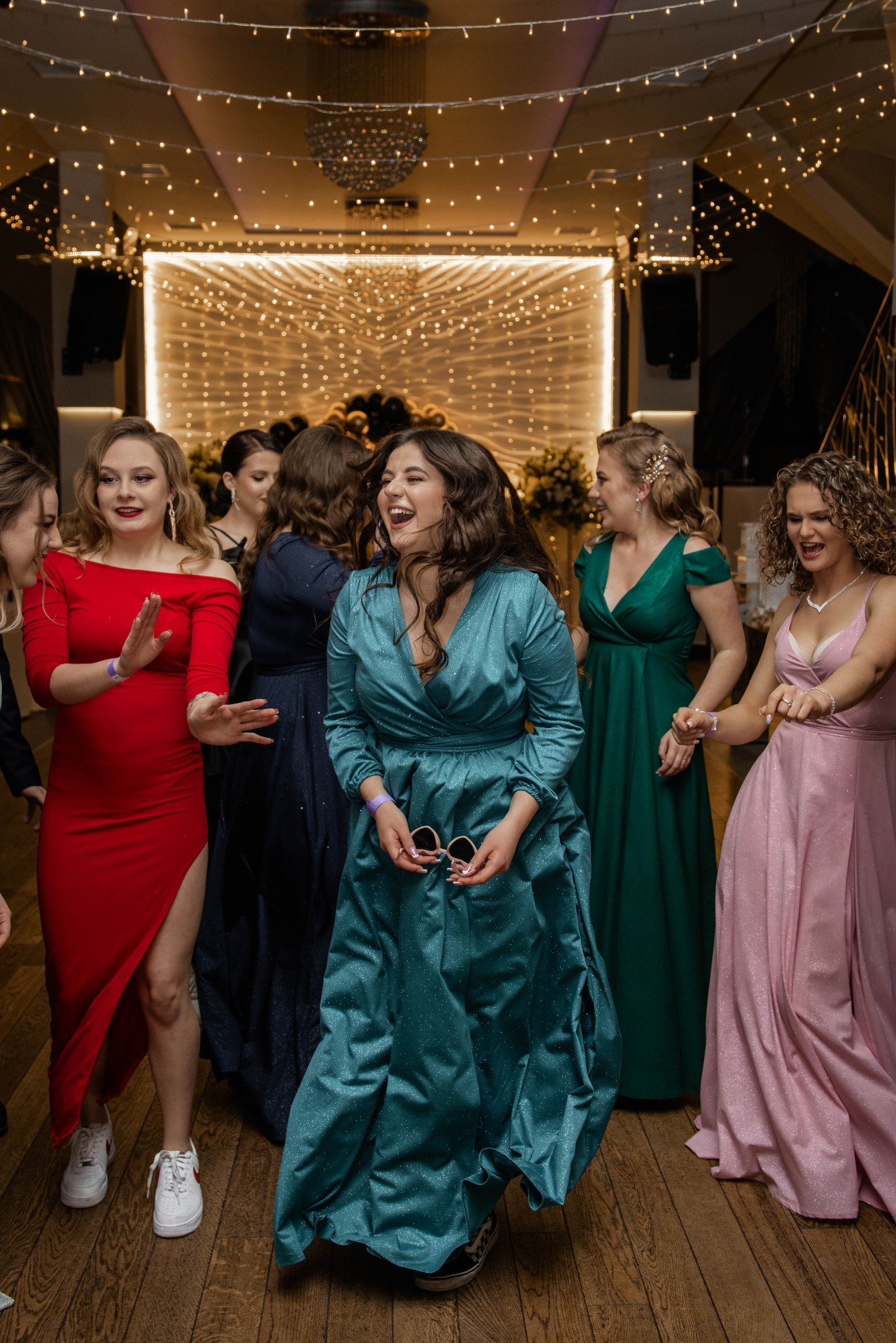 Grupa roześmianych kobiet w sukniach wieczorowych tańczy na parkiecie podczas imprezy, widoczne dekoracje świetlne i żyrandol.