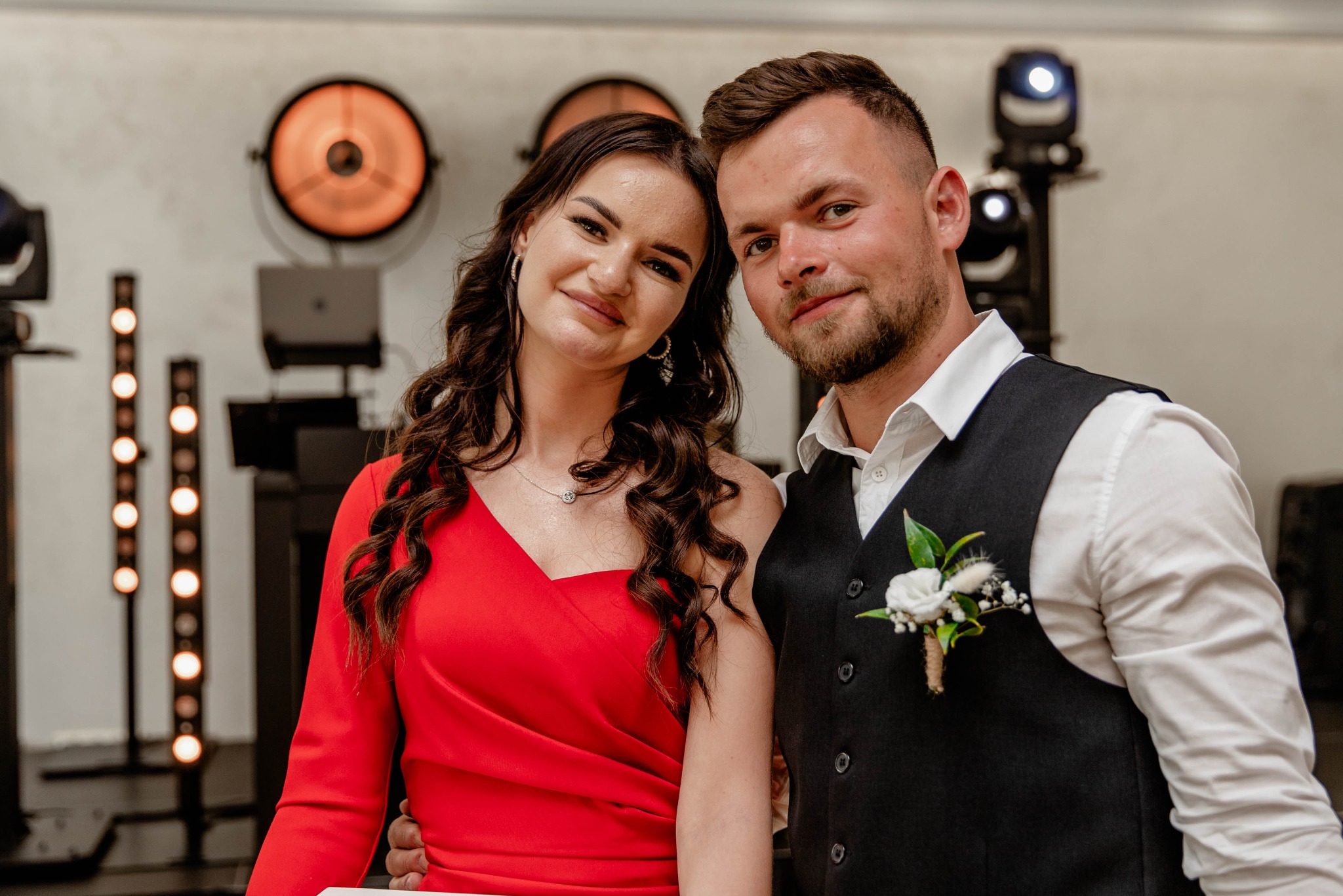 Portret uśmiechniętej pary: kobieta w czerwonej sukience i mężczyzna w czarnej kamizelce z butonierką, na tle dekoracji świetlnych.