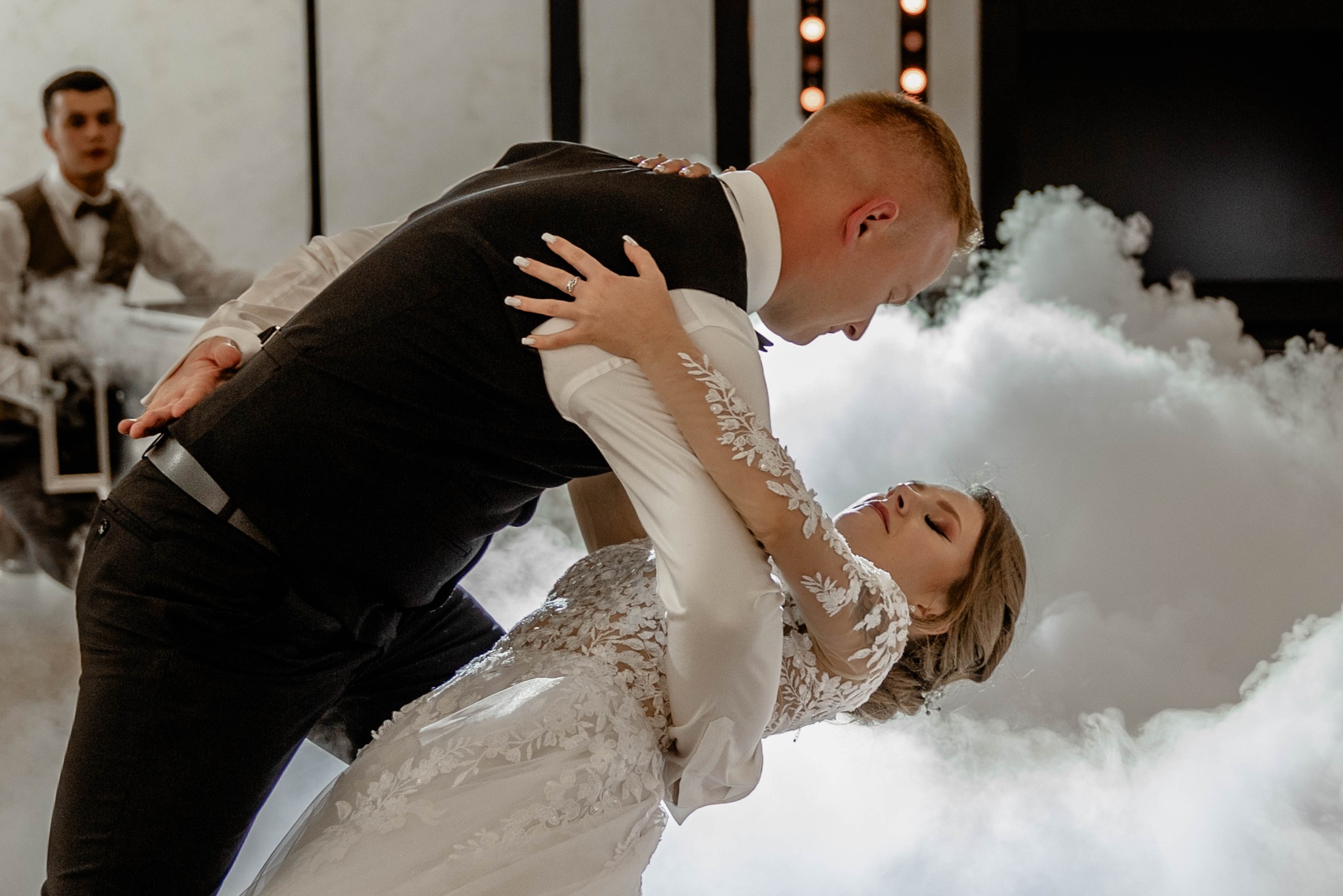 Para młoda w tańcu, panna młoda w koronkowej sukni z długimi rękawami, pan młody w czarnym garniturze, tło wypełnione efektem dymu i światłami, w tle obserwujący mężczyzna.