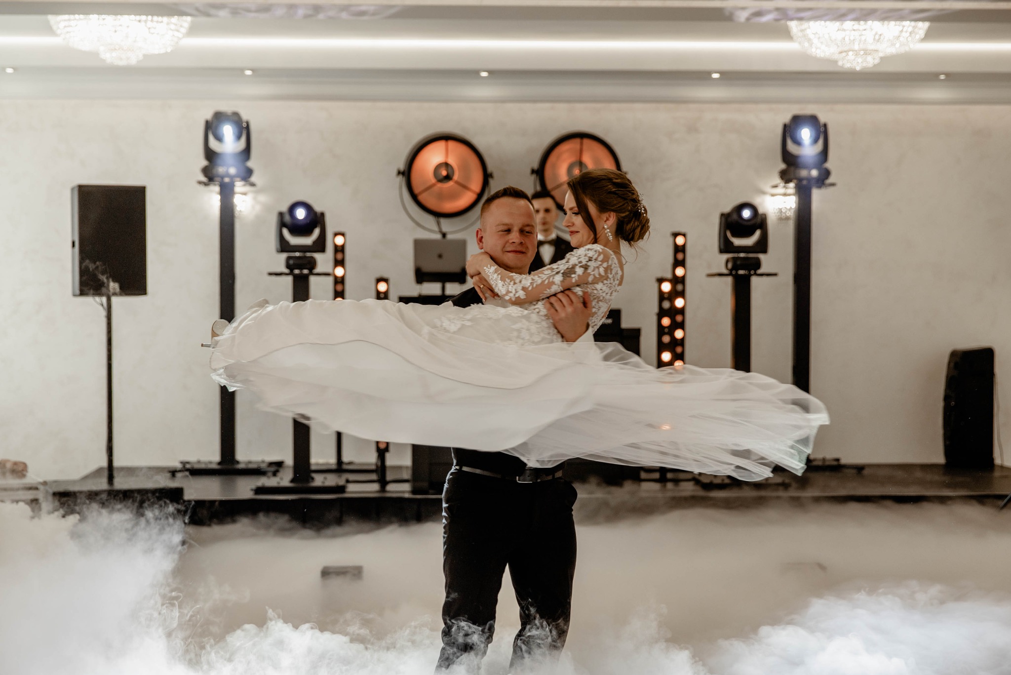 Pan młody trzyma pannę młodą w ramionach na parkiecie w chmurze dymu, w tle oświetlenie sceniczne.