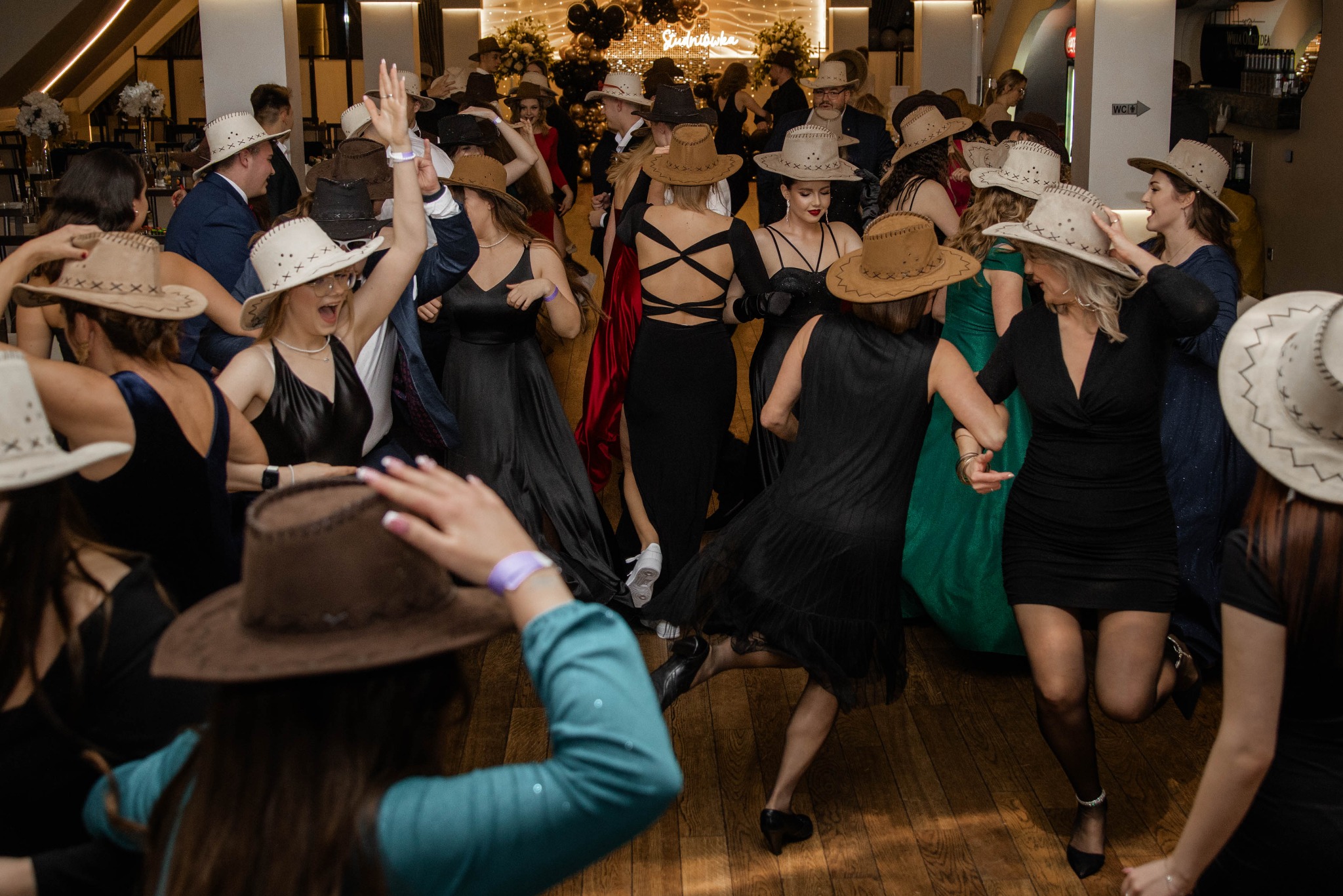Rozbawieni goście w kowbojskich kapeluszach tańczą w sali balowej podczas studniówki, widoczny napis 'Studniówka' w tle.