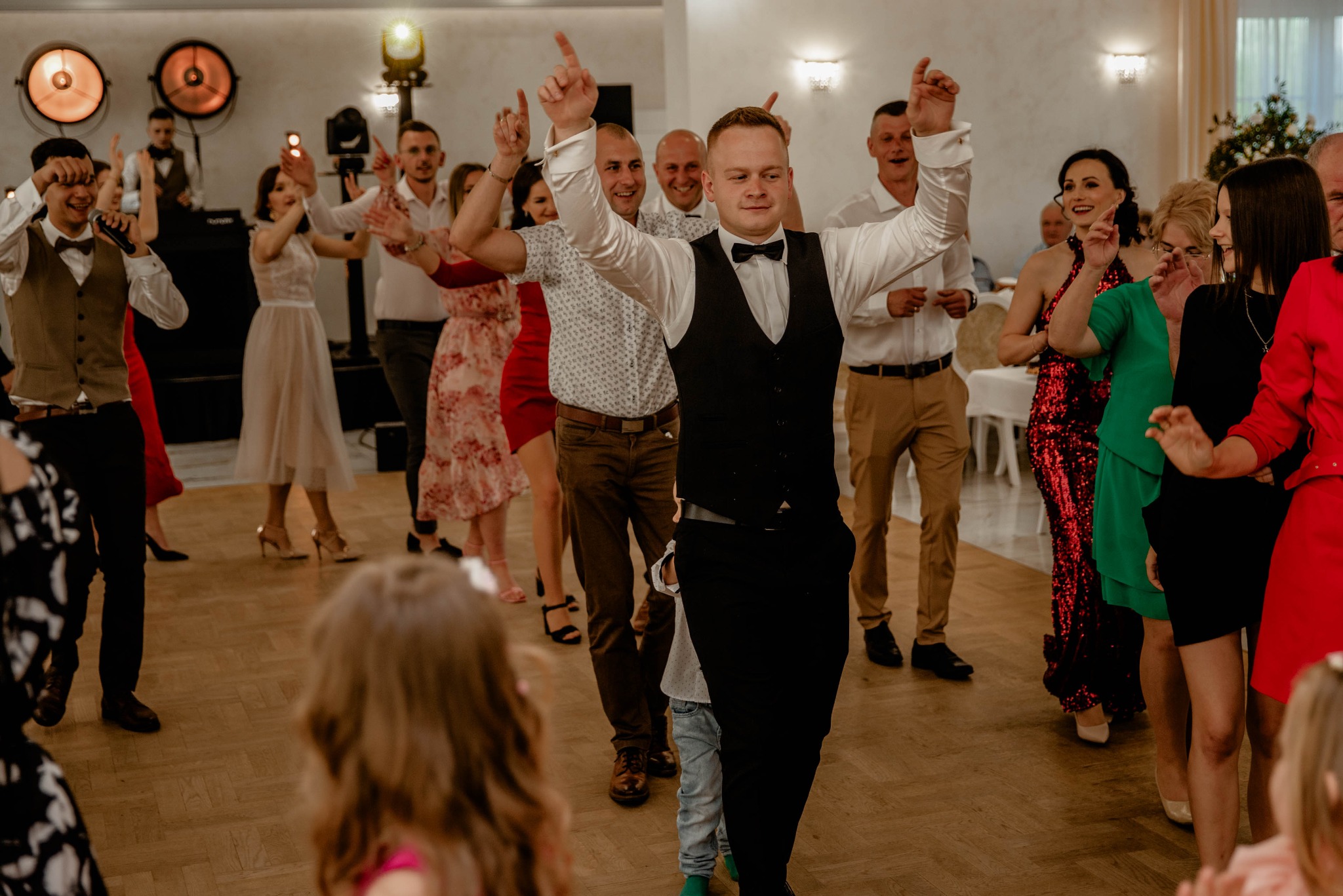 Goście weselni tańczący na parkiecie, w tle DJ z mikrofonem, oświetlenie punktowe