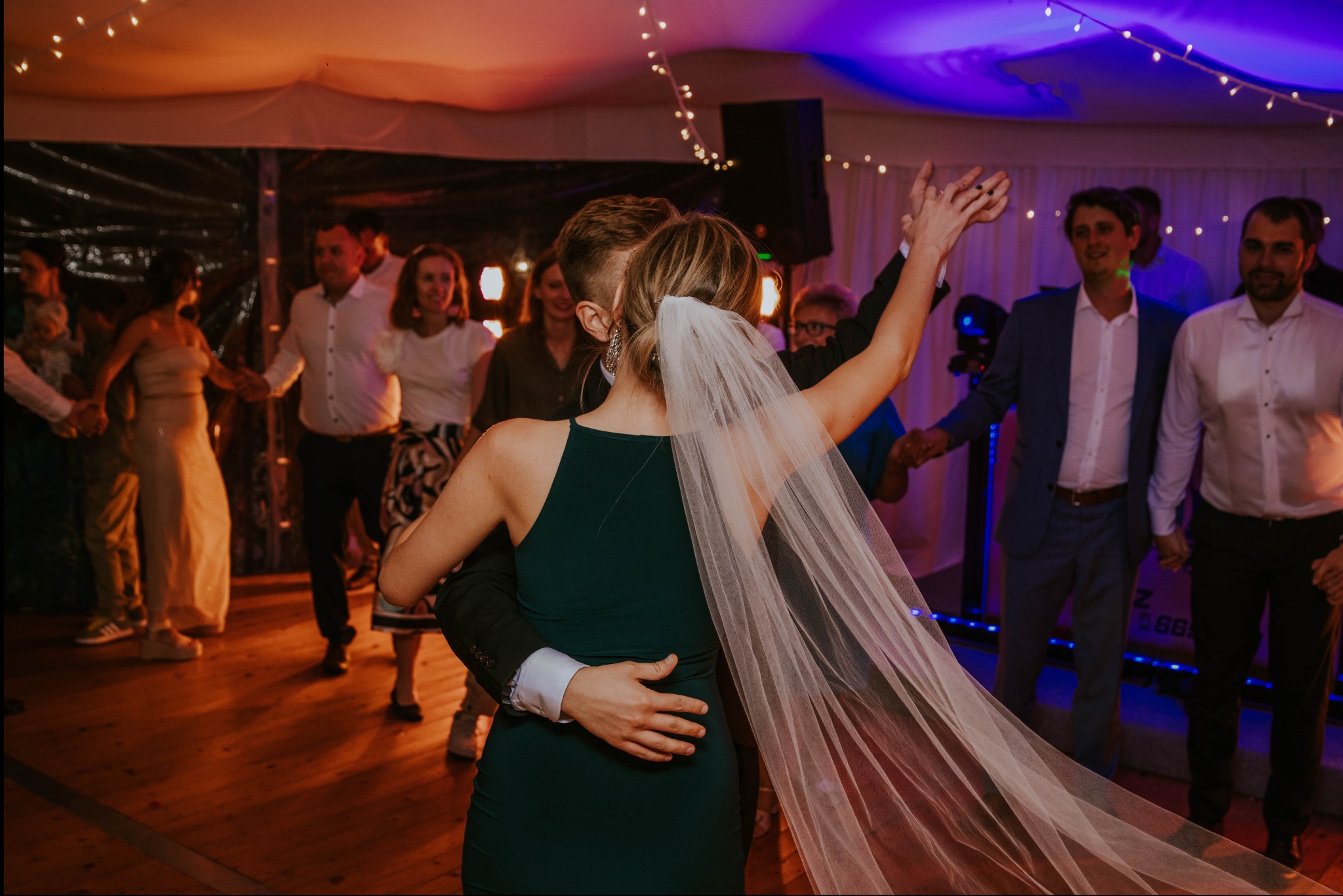 Para młoda tańczy podczas wesela, panna młoda w zielonej sukience i welonie, goście weselni w tle, oświetlenie sceniczne i girlandy świetlne.