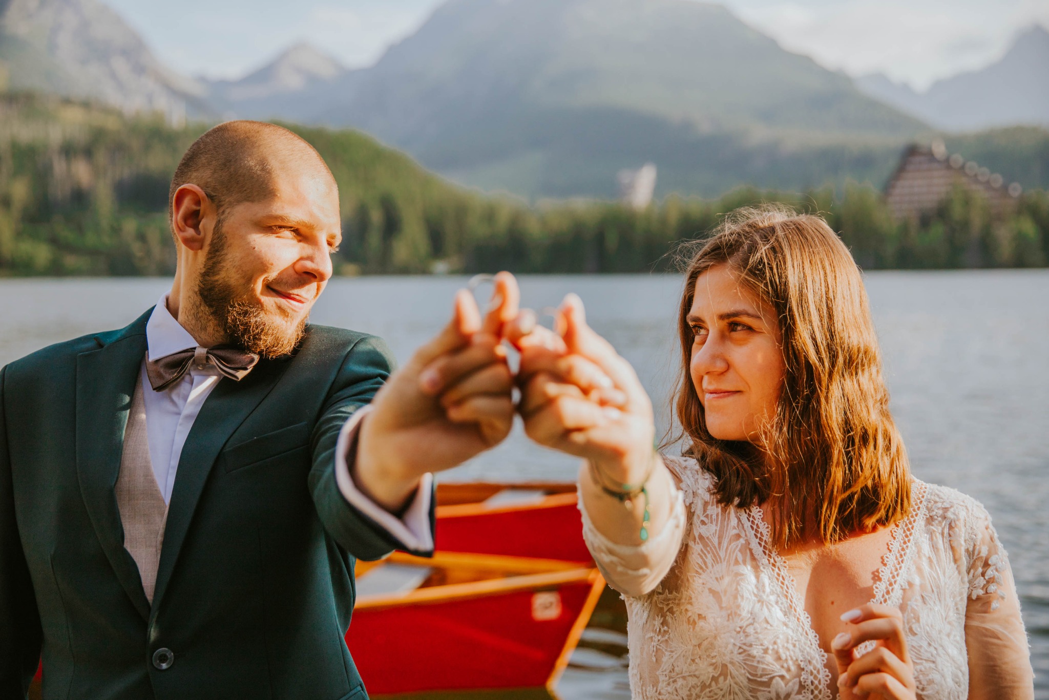 Para młoda trzymająca dłonie z pierścionkami na tle jeziora i gór, on w zielonym garniturze i muszce, ona w białej koronkowej sukience.
