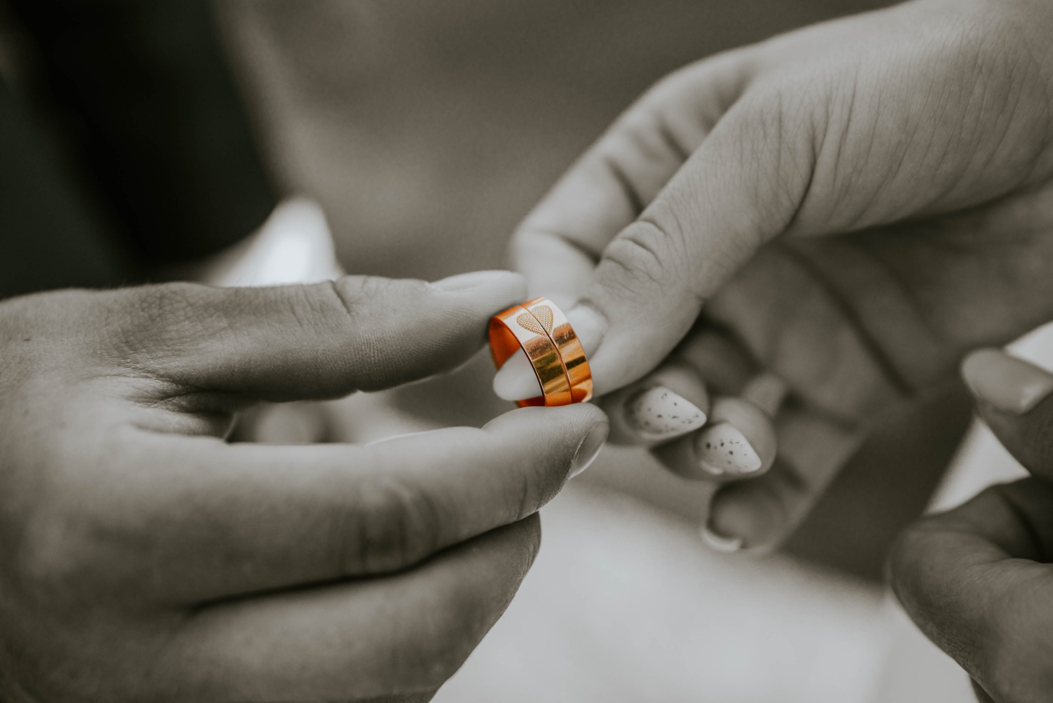 Dwie ręce trzymające złote obrączki z wygrawerowanym wzorem serca. Fotografia w odcieniach szarości z akcentem na kolor obrączek.