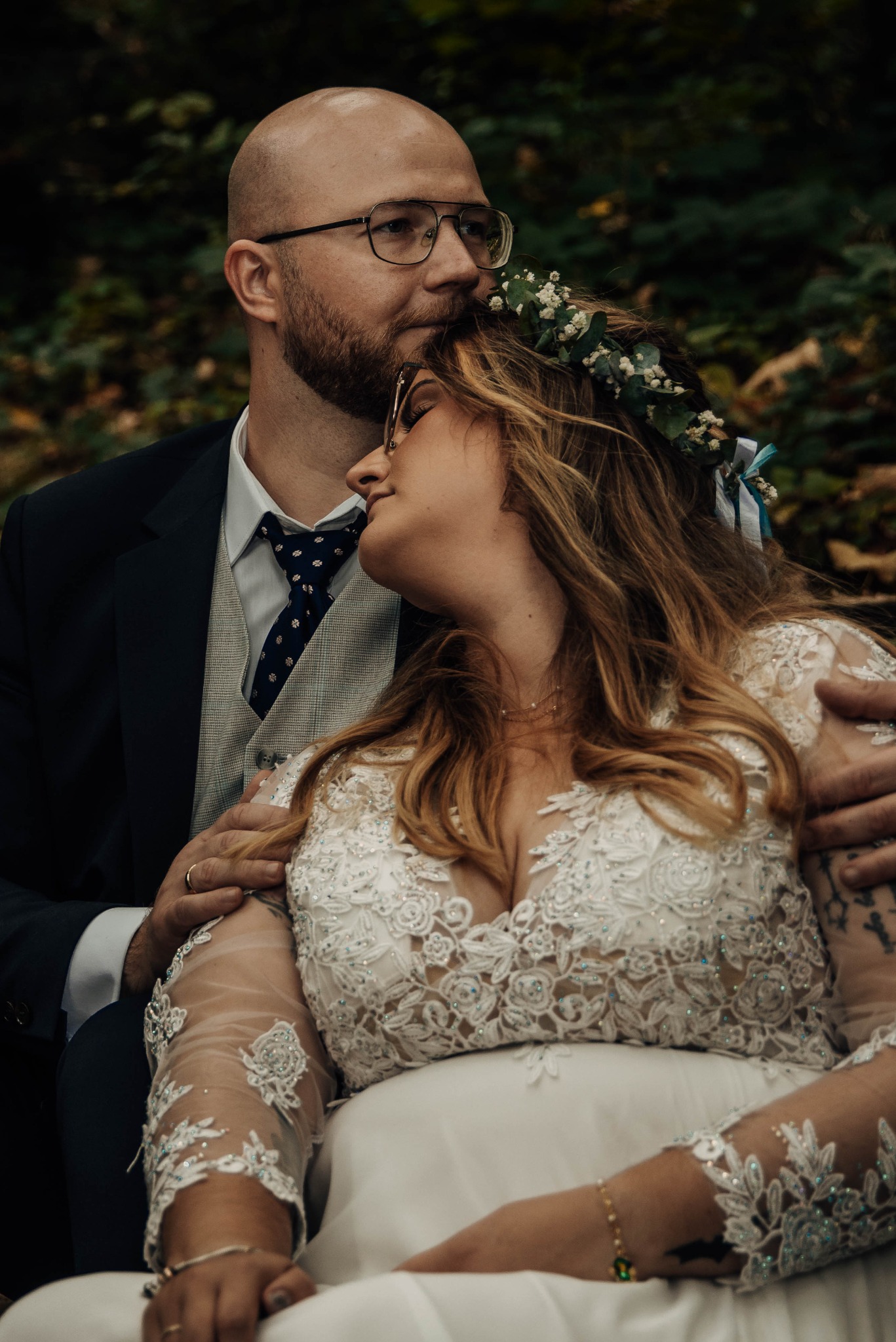Para młoda w objęciach na tle zieleni: panna młoda w koronie z kwiatów i sukni z koronkowymi aplikacjami, pan młody w garniturze i okularach.