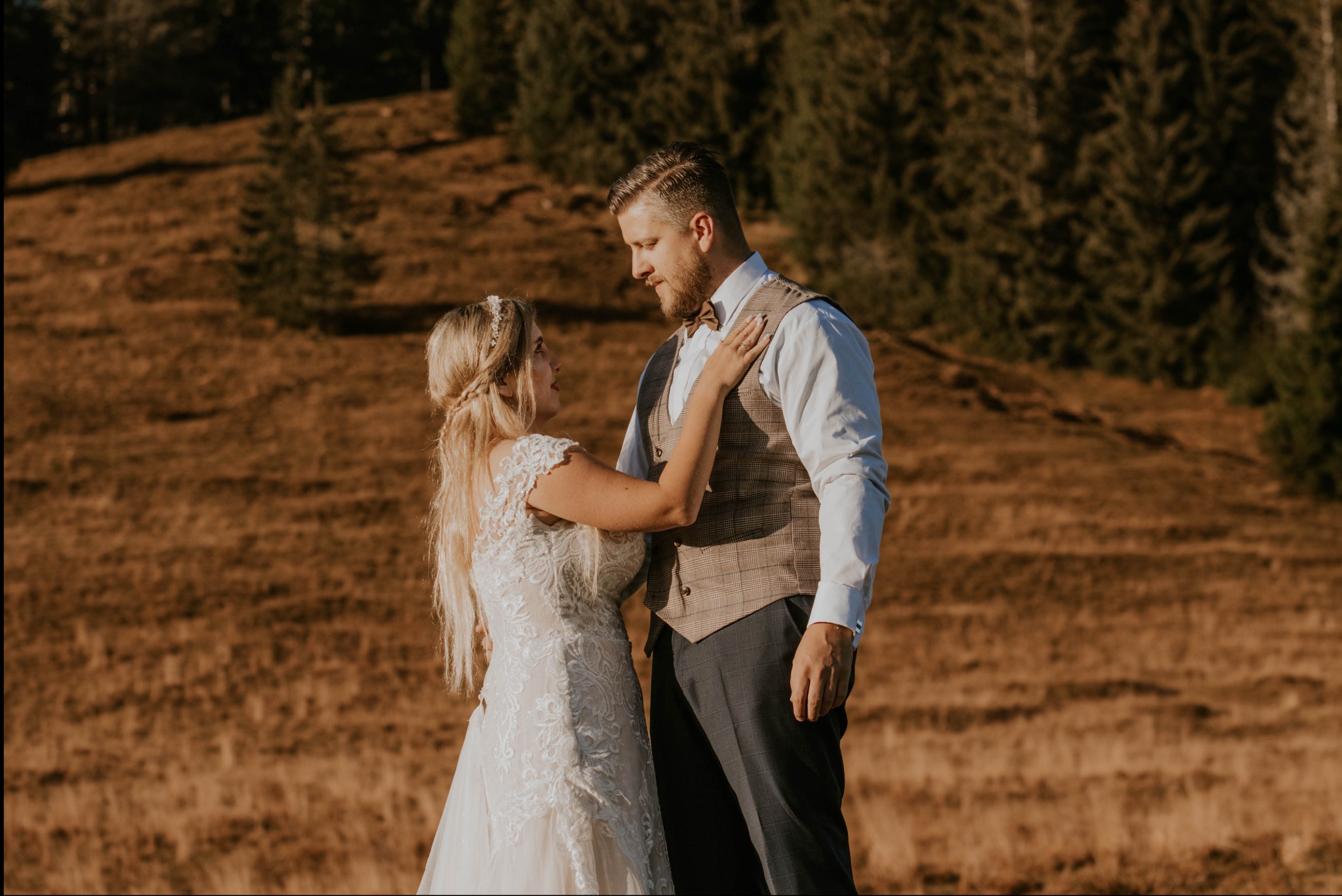 Para młoda w plenerze: panna młoda w koronkowej sukni z warkoczem i ozdobą we włosach, dotyka ramienia pana młodego w brązowej kamizelce w kratę i muszce, na tle wzgórza z drzewami.