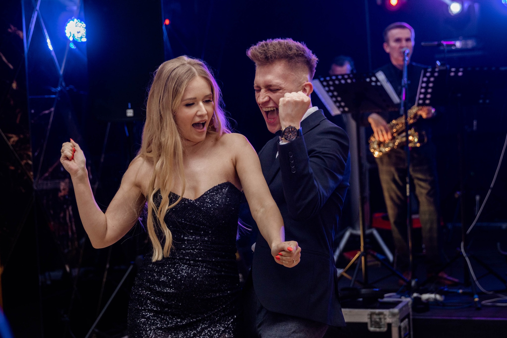 Para młoda w tańcu podczas wesela, w tle zespół muzyczny z saksofonem, dynamiczne oświetlenie sceniczne.