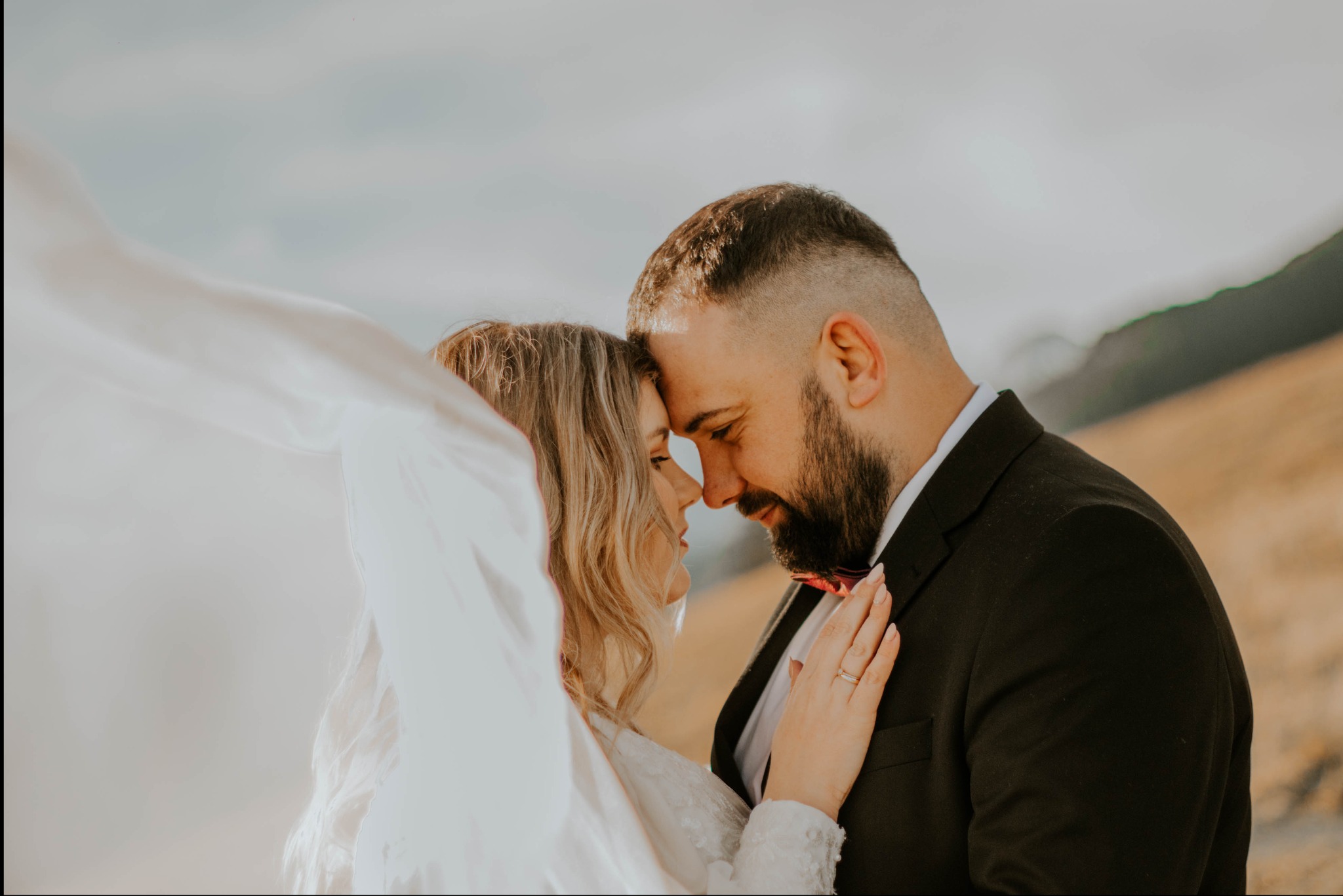 Para młoda dotykająca się czołami na tle górskim, welon powiewa na wietrze, on w czarnym garniturze z bordową muchą, ona w białej sukni z koronkowymi rękawami i delikatnym makijażem.