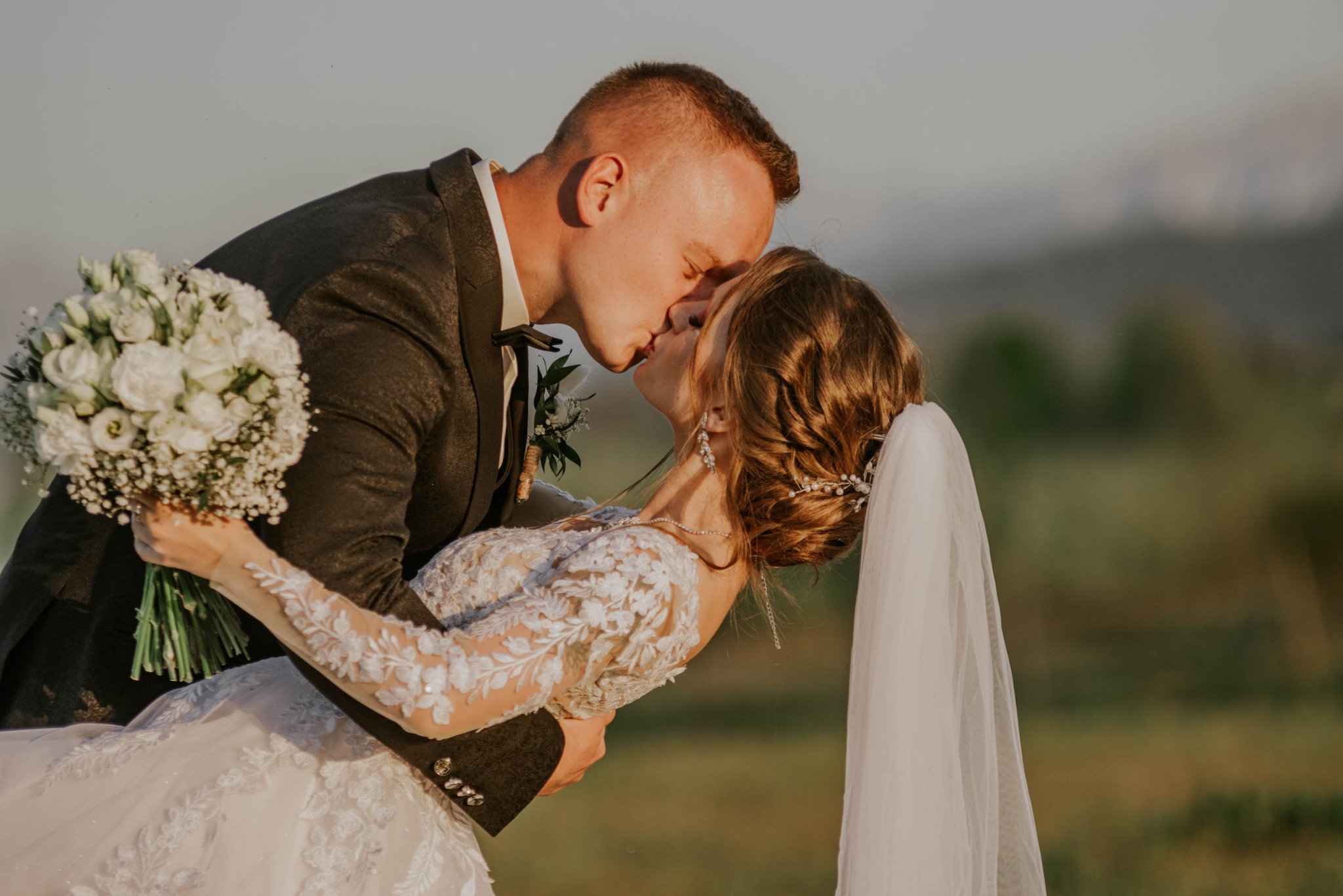 Pocałunek nowożeńców w plenerze: panna młoda w koronkowej sukni z welonem, trzymająca bukiet białych kwiatów, pochylona w ramionach pana młodego w ciemnym garniturze.
