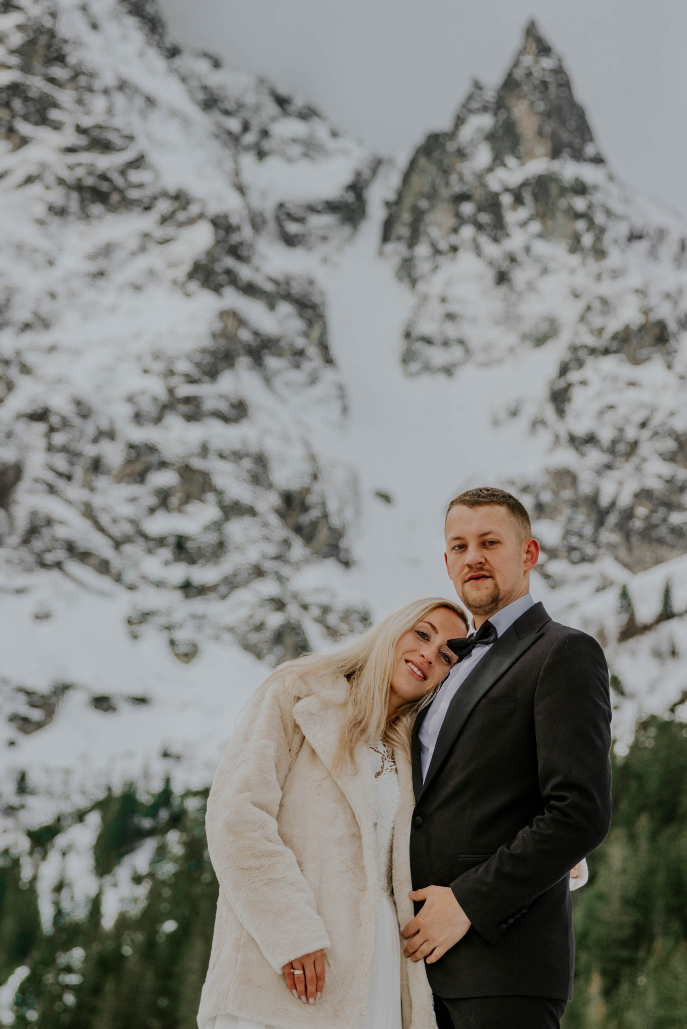Para młoda: blondynka w futrze opiera głowę na ramieniu mężczyzny w czarnym smokingu na tle ośnieżonych gór.