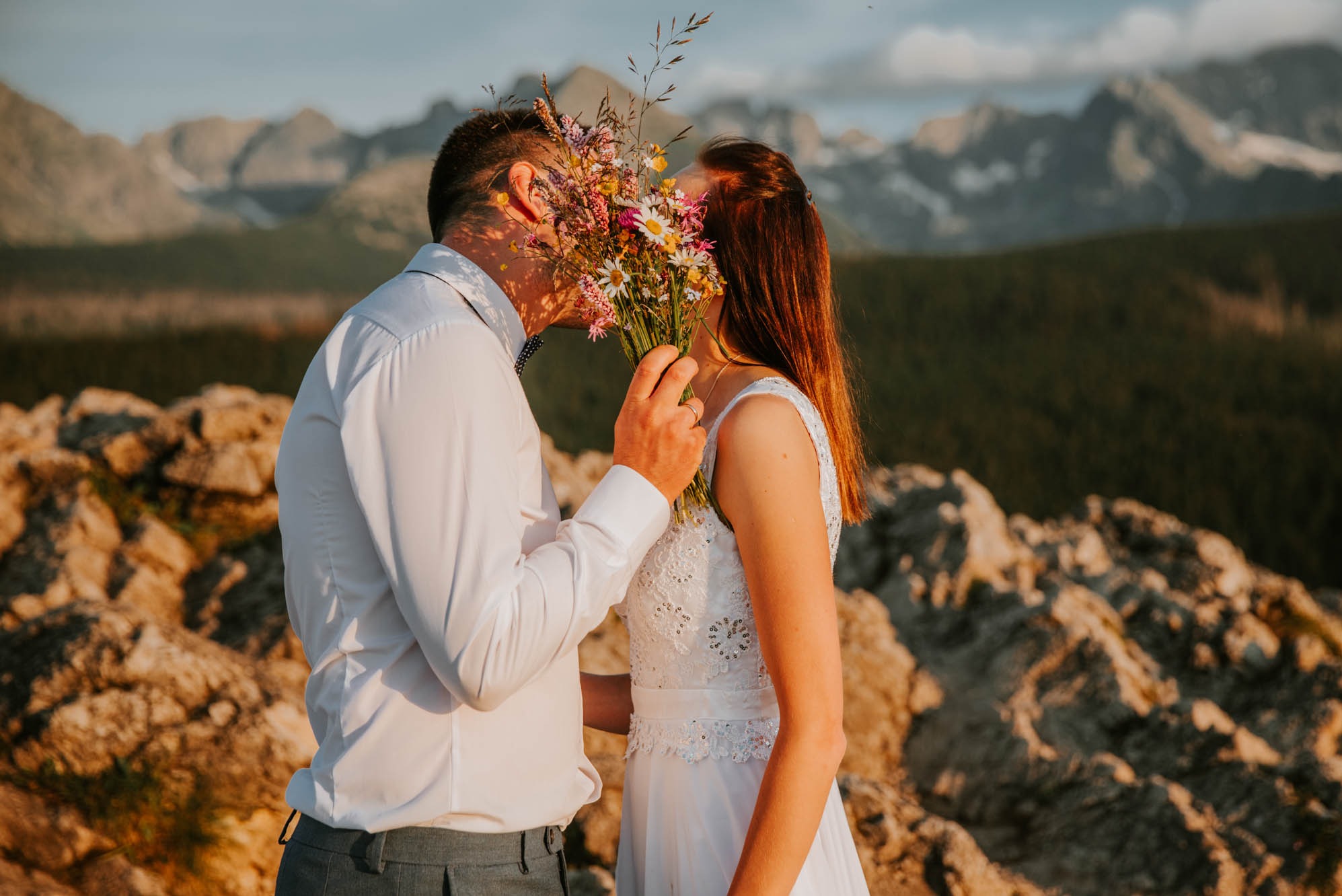 Para młoda całuje się za bukietem polnych kwiatów na tle górskiego krajobrazu o zachodzie słońca. Panna młoda w białej sukni z koronkowymi detalami, pan młody w białej koszuli i szarych spodniach.