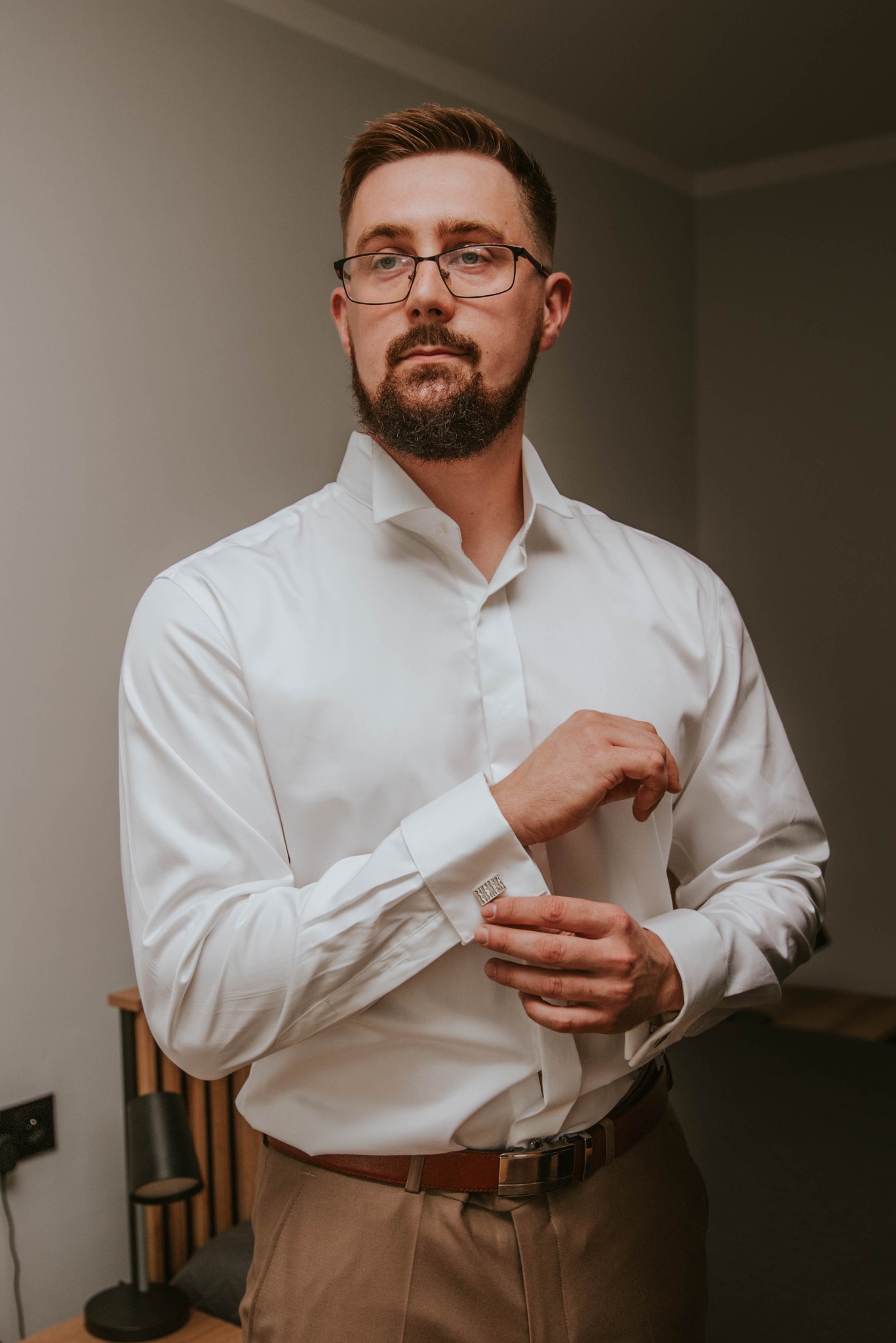 Mężczyzna w okularach, zapinający mankiety koszuli z ozdobnymi spinkami, ubrany w białą koszulę i brązowe spodnie, przygotowuje się do wyjścia.