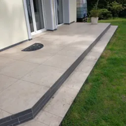 Wykończony taras z beżowych płytek z ciemnymi obrzeżami i schodami, widok z poziomu trawnika. Na tarasie wycieraczka i donica z rośliną. W tle ściana budynku i zieleń.