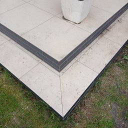 ARR-BUD - Wykończone schody zewnętrzne z jasnych płytek i ciemnoszarego obrzeża, prowadzące do tarasu z donicą. Widok z góry na połączenie płytek i trawy.