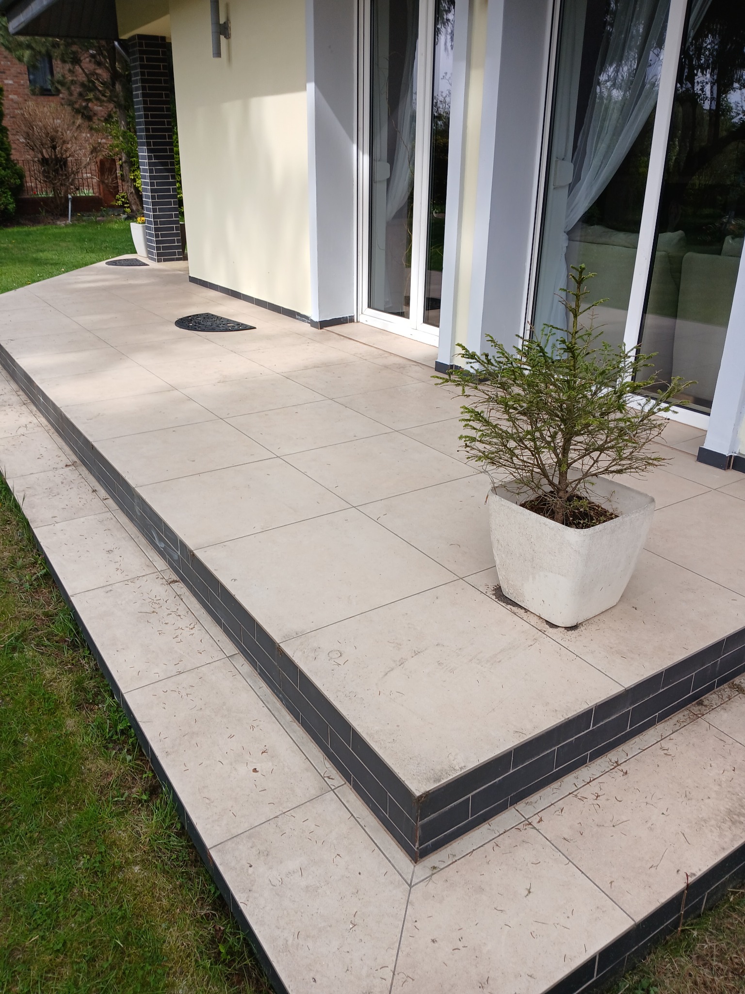 Jasne płytki tarasowe z ciemną obwódką na schodach, donica z iglakiem, przeszklone drzwi tarasowe. Widok na zadbany trawnik i elewację domu.