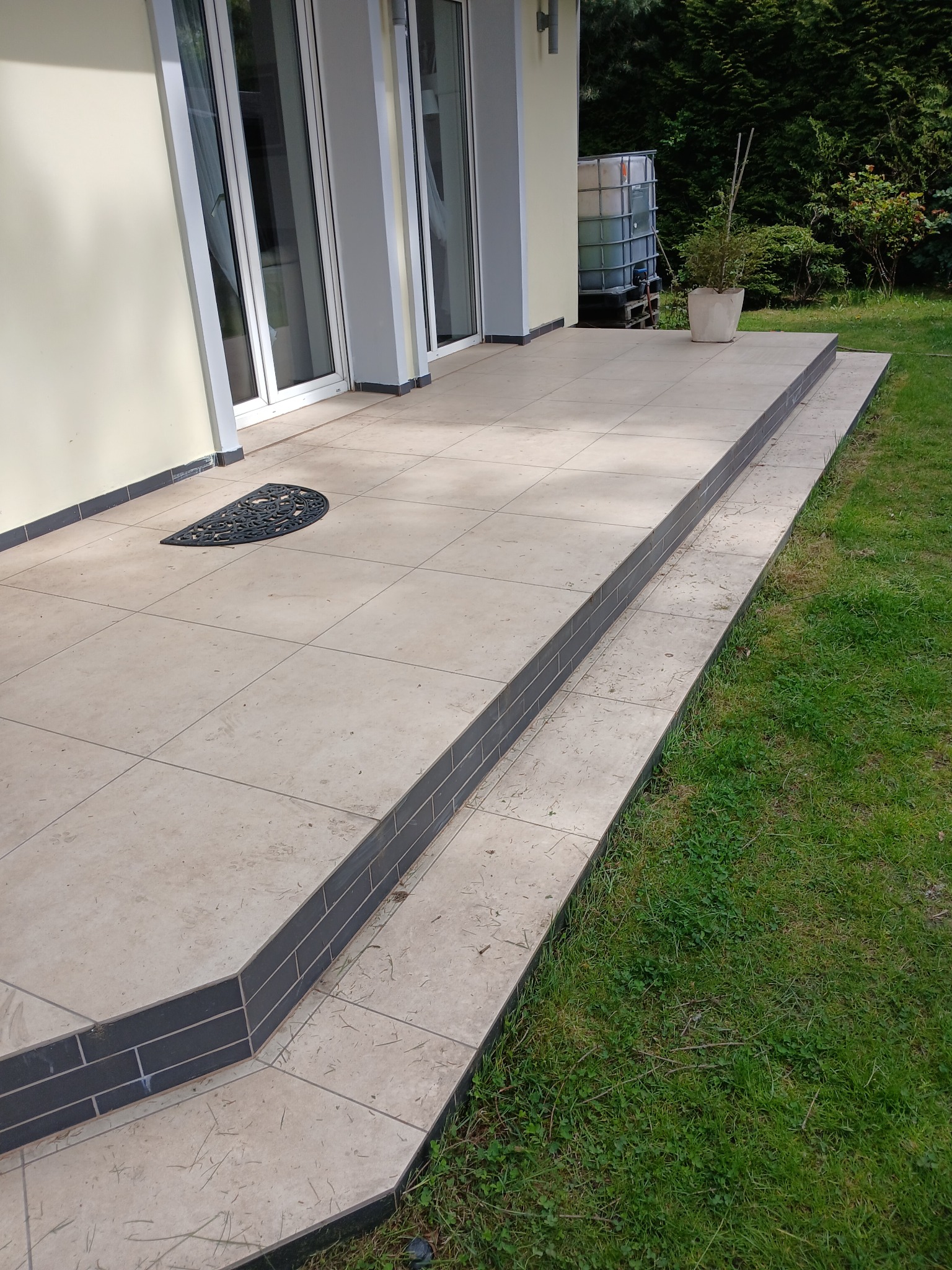 Wykończony taras z beżowych płytek z ciemnymi obrzeżami i schodami, widok z poziomu trawnika. Na tarasie wycieraczka i donica z rośliną. W tle ściana budynku i zieleń.