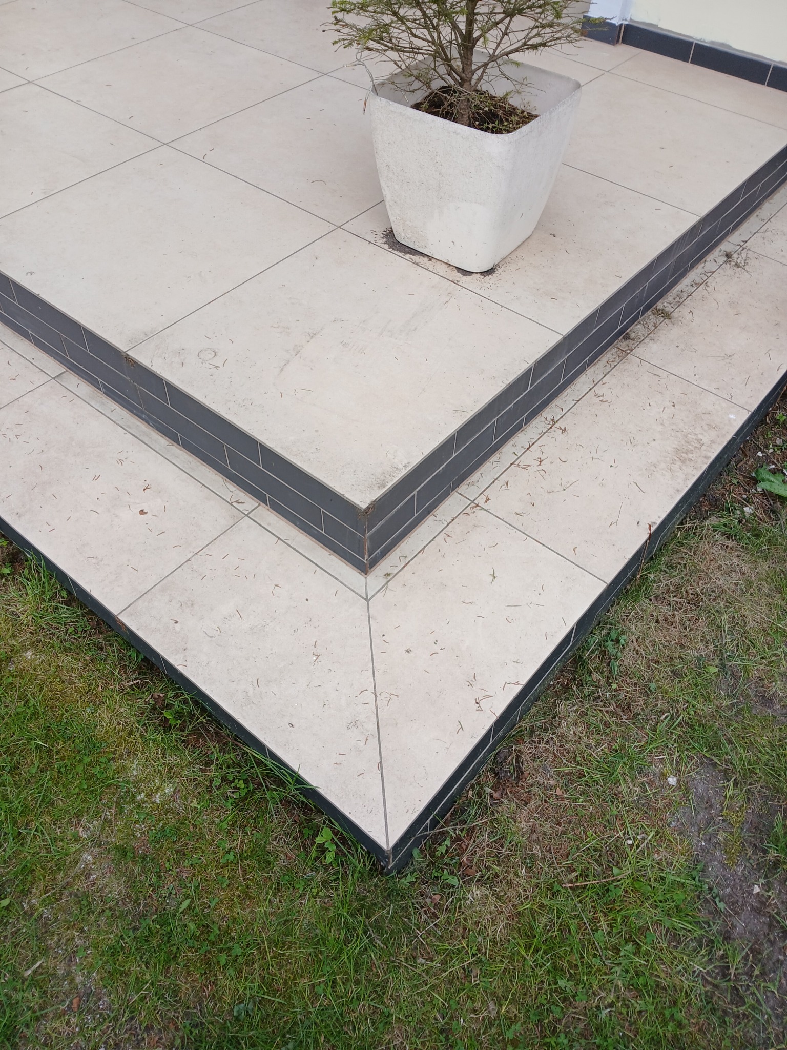 Wykończone schody zewnętrzne z jasnych płytek i ciemnoszarego obrzeża, prowadzące do tarasu z donicą. Widok z góry na połączenie płytek i trawy.