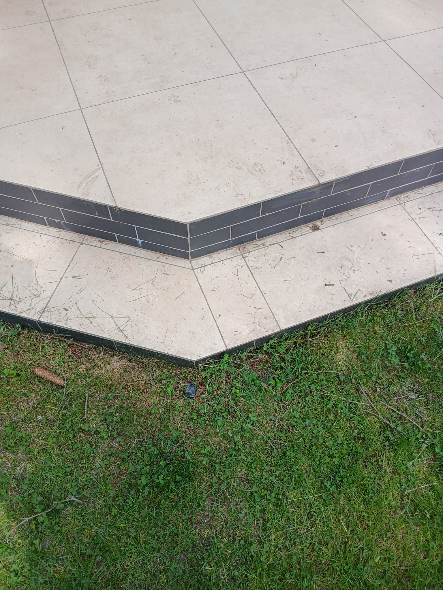 Wykończone płytkami schody tarasowe z ciemnym obramowaniem, przechodzące w trawnik. Widoczne ślady użytkowania na jasnych płytkach. Kątowe ujęcie.