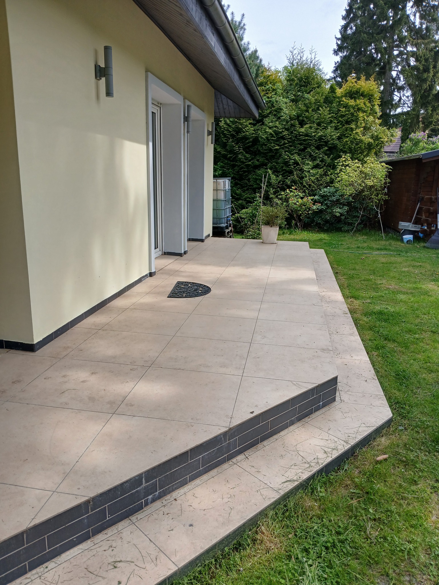 Wyłożony beżowymi płytkami taras z dwoma stopniami, obok trawnik i ściana budynku z lampami. Na tarasie wycieraczka półokrągła i doniczka z rośliną.