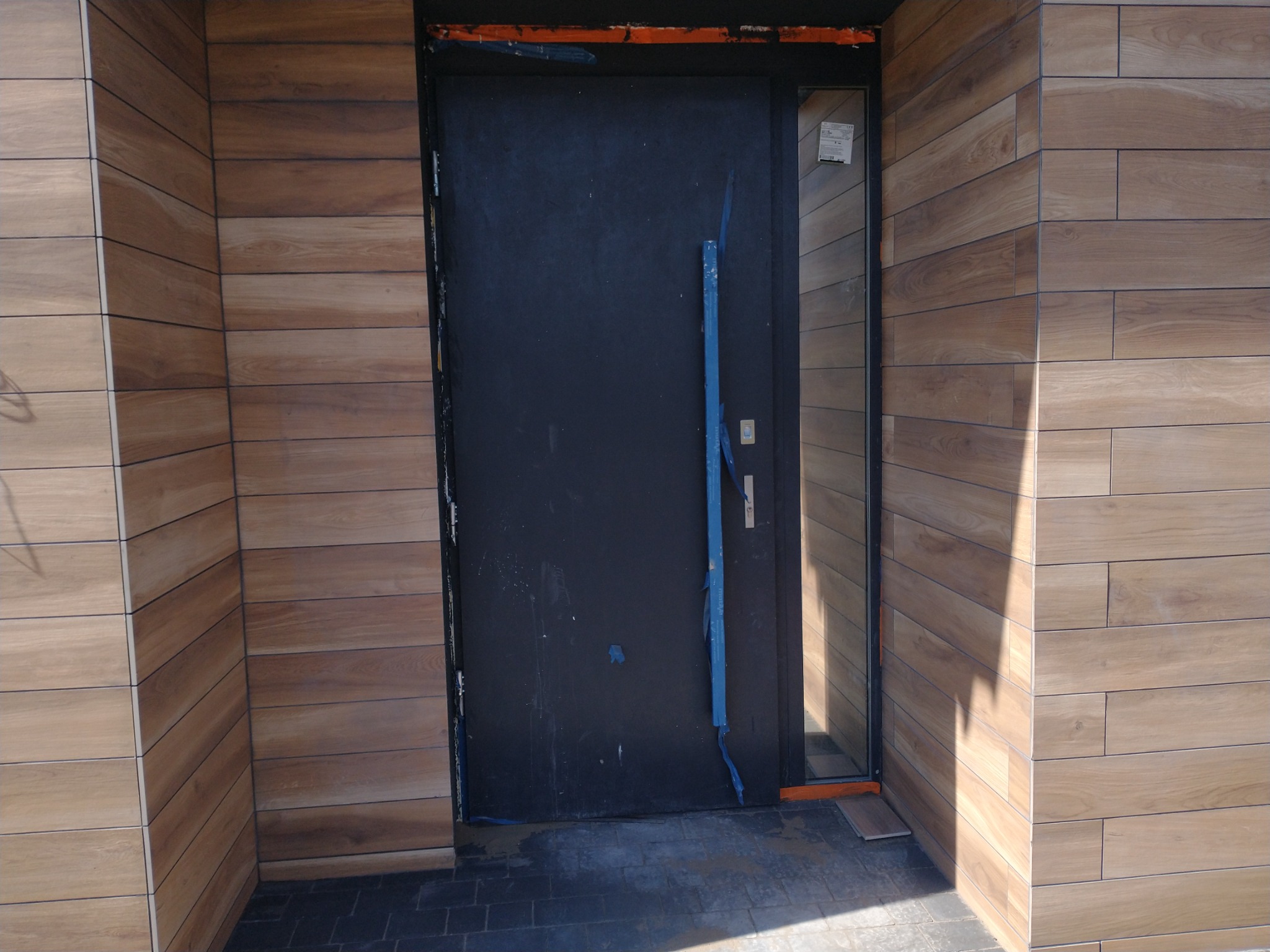 Wejście do budynku z drewnopodobną okładziną elewacyjną i czarnymi drzwiami, zabezpieczonymi folią. Detal wykończenia zewnętrznego.