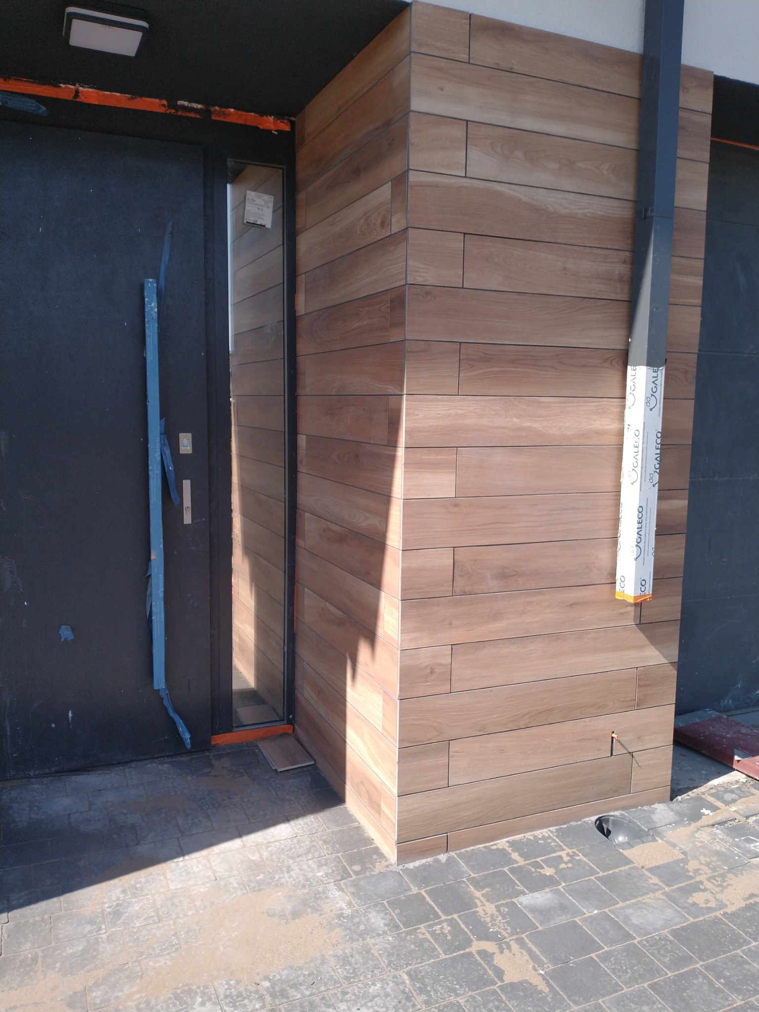 Wykończenie elewacji płytkami drewnopodobnymi przy wejściu do budynku. Widoczna czarna stolarka drzwiowa i szara kostka brukowa.