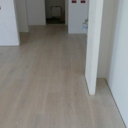 Paweł Mroszczyk - Jasne panele podłogowe ułożone w nowym mieszkaniu, widok z perspektywy podłogi, białe ściany i przygotowane otwory na instalacje elektryczne w tle.